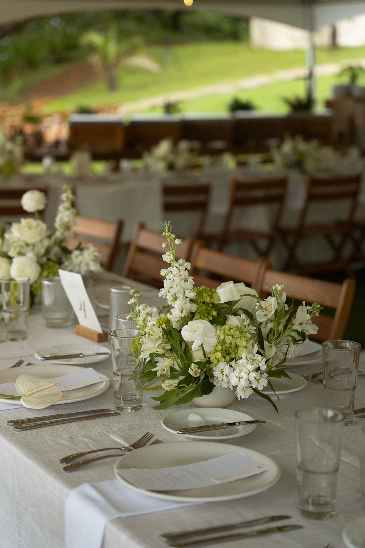 A long reception table styled with white linens, wooden chairs, and soft floral centerpieces beneath an open tent at one of the most elegant Kauai wedding venues.