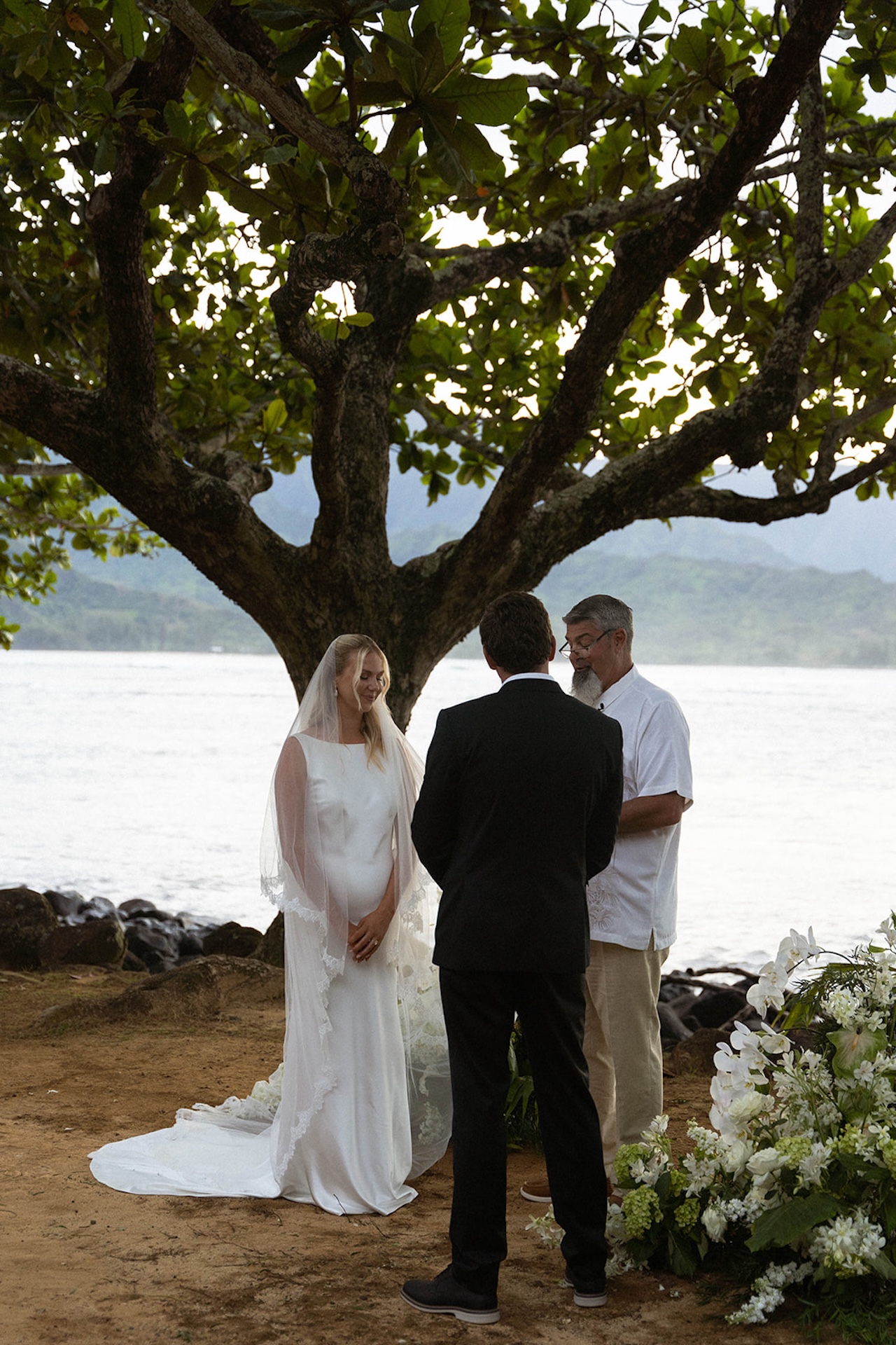 A bride and groom stand facing their officiant beneath a large tree by the ocean at one of the most intimate Kauai wedding venues.