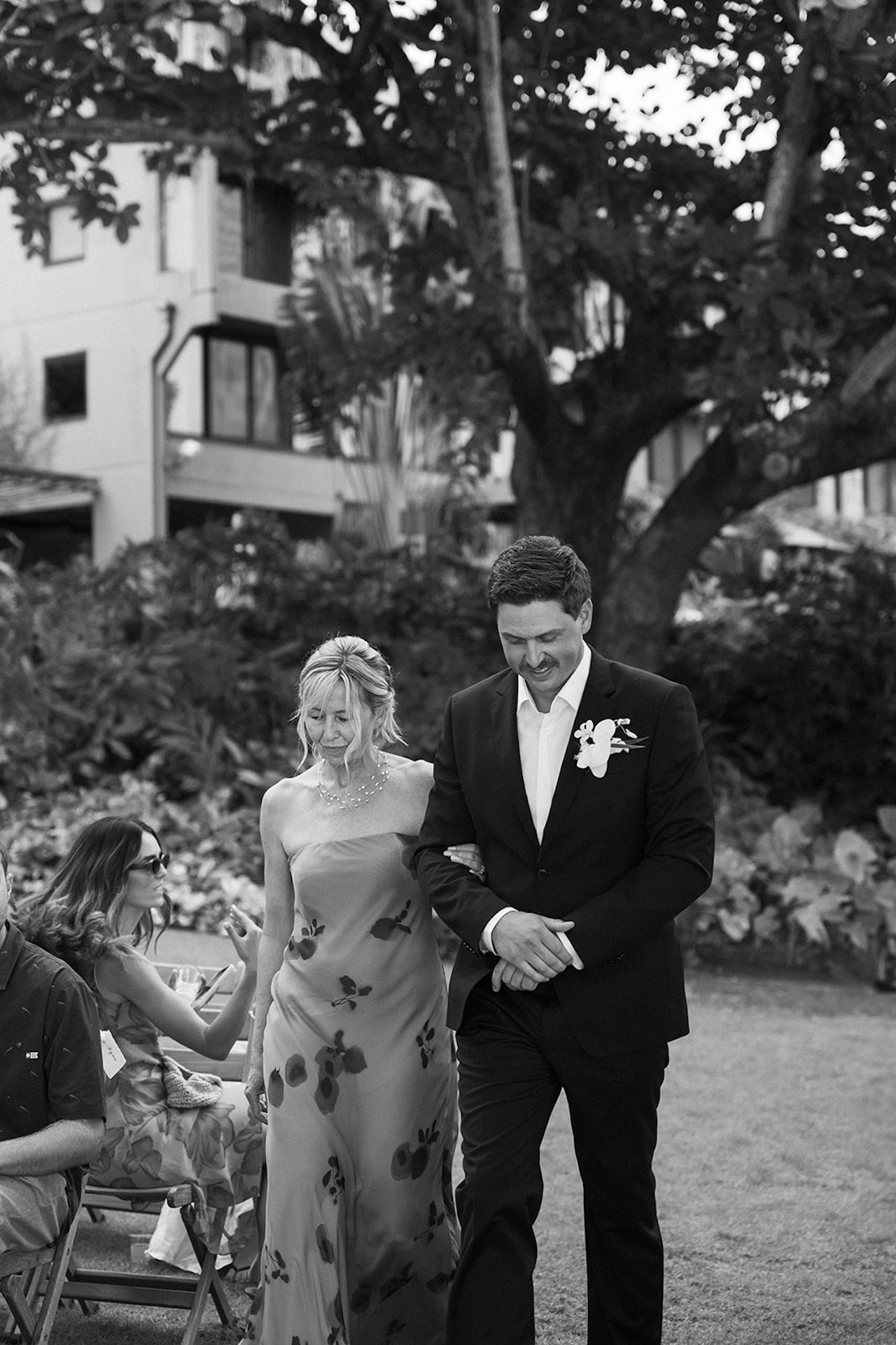 Black and white image of a groom walking arm in arm with his mom, capturing candid wedding moments during an outdoor reception.