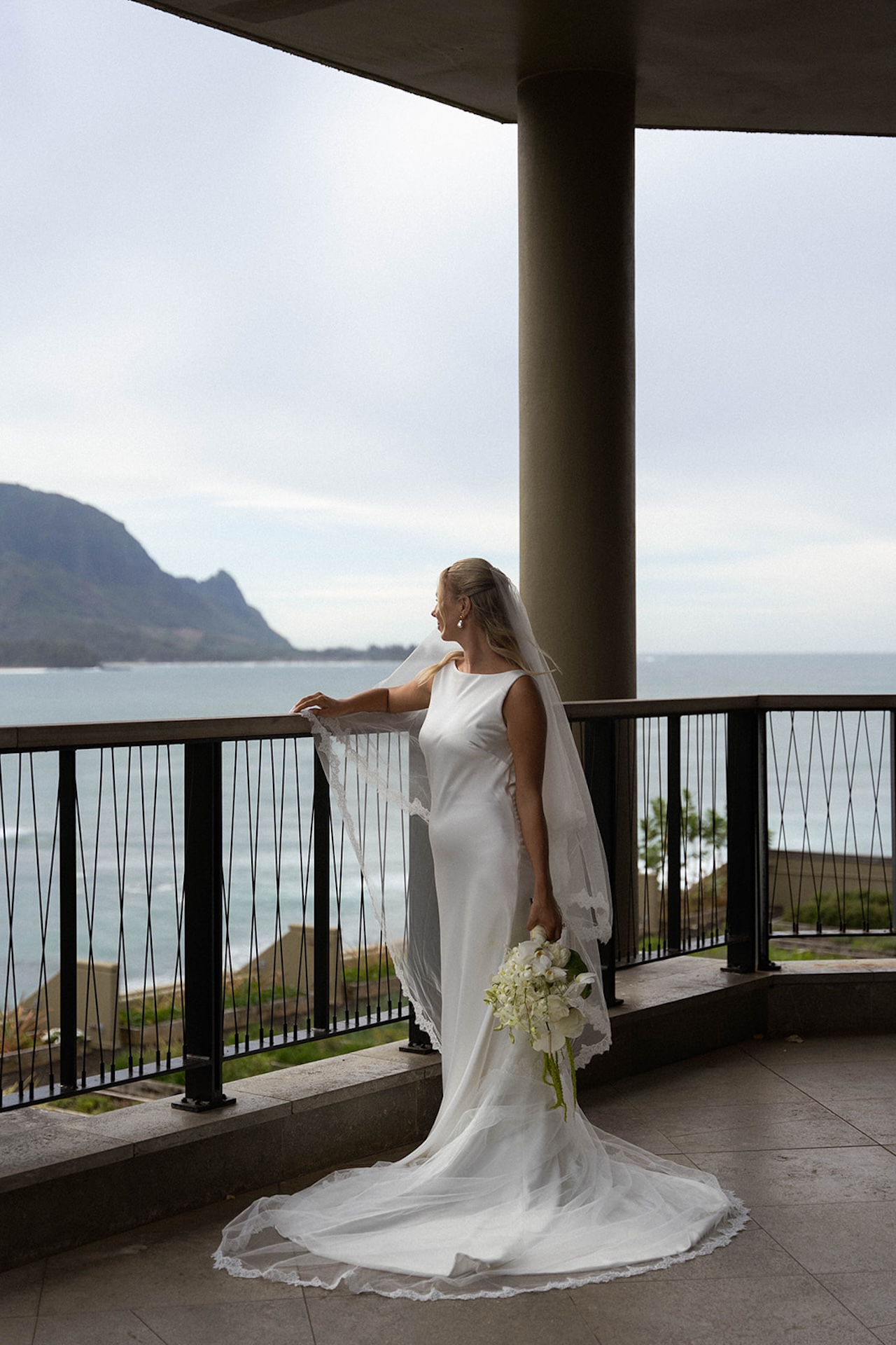 A bride gazes out over the ocean from a covered terrace, wearing a sleek wedding gown and veil with a coastal mountain backdrop.