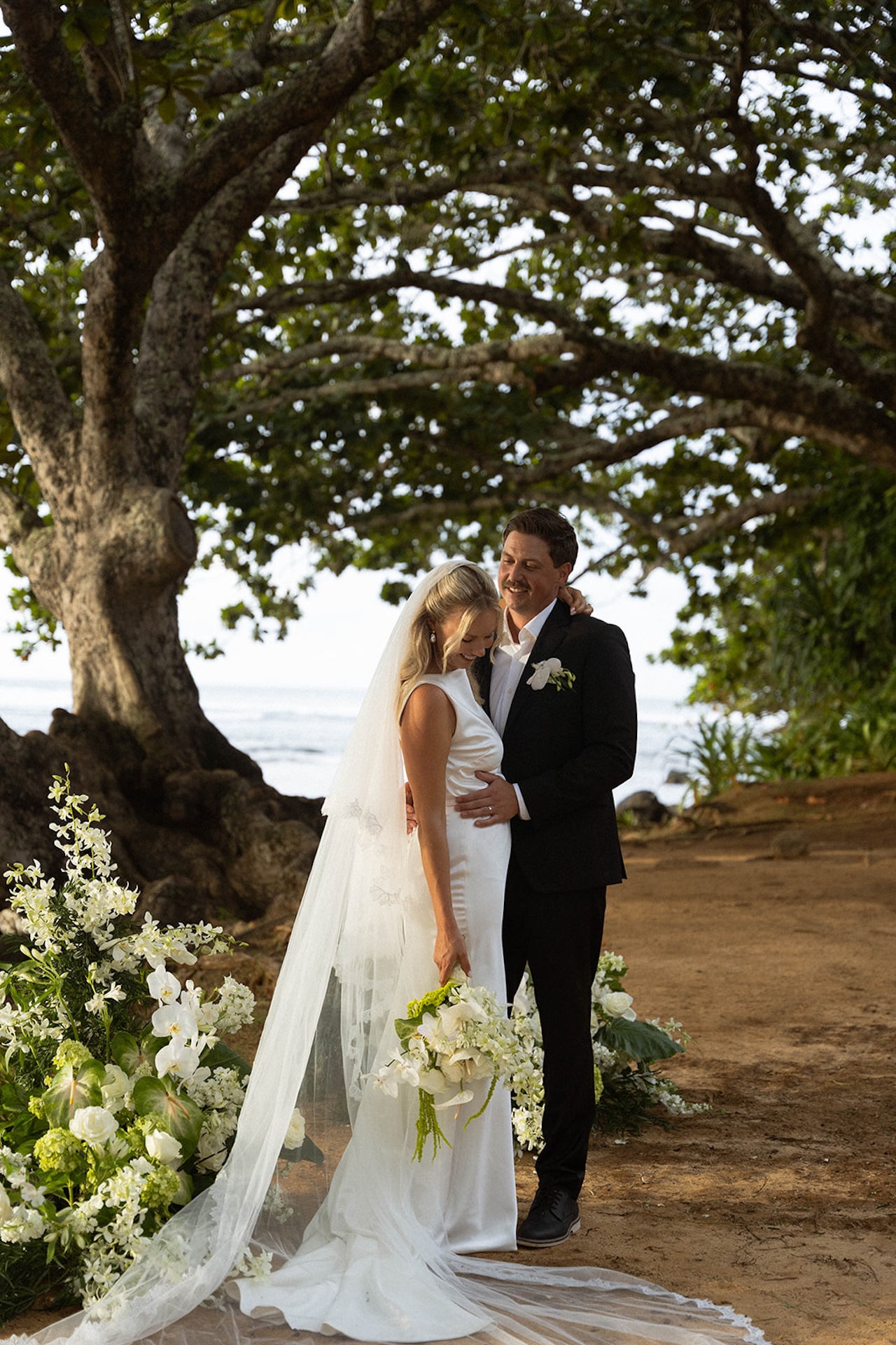 A newly married couple embraces beneath tropical trees with the coastline behind them at one of the most romantic Kauai wedding venues.