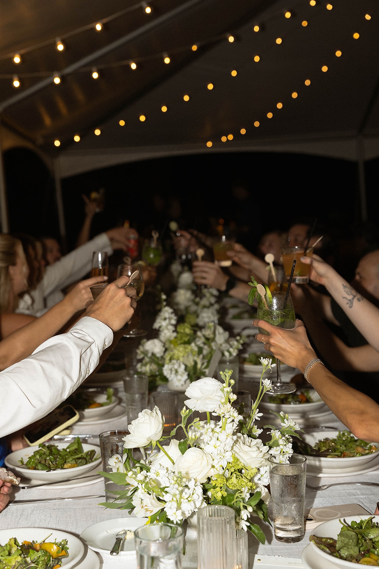 Guests raise their glasses for a lively toast beneath a sailcloth tent with string lights glowing above the reception table.