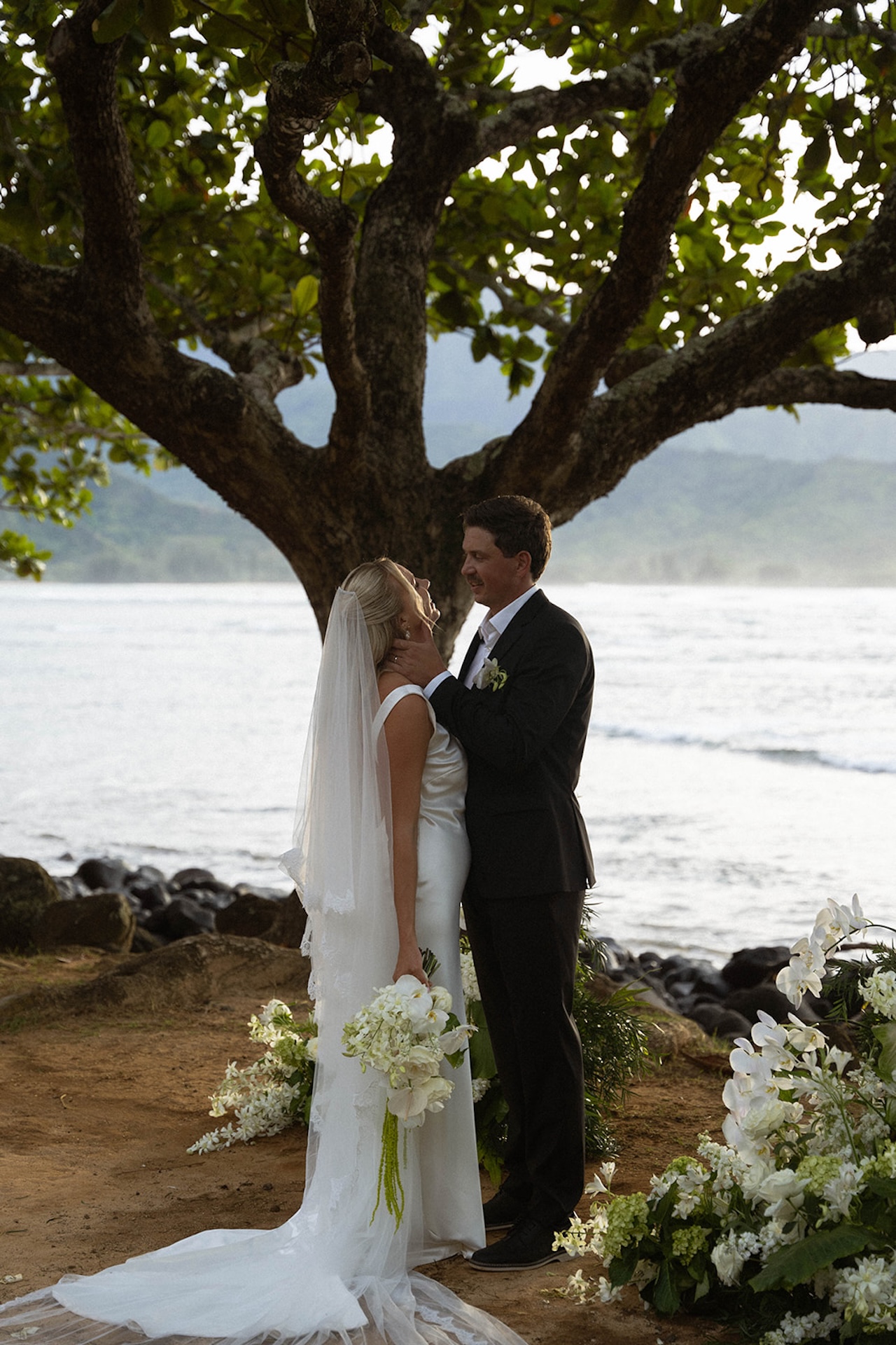 The bride and groom share a quiet moment beneath a tree by the ocean, framed by florals at one of the most intimate Kauai wedding venues.