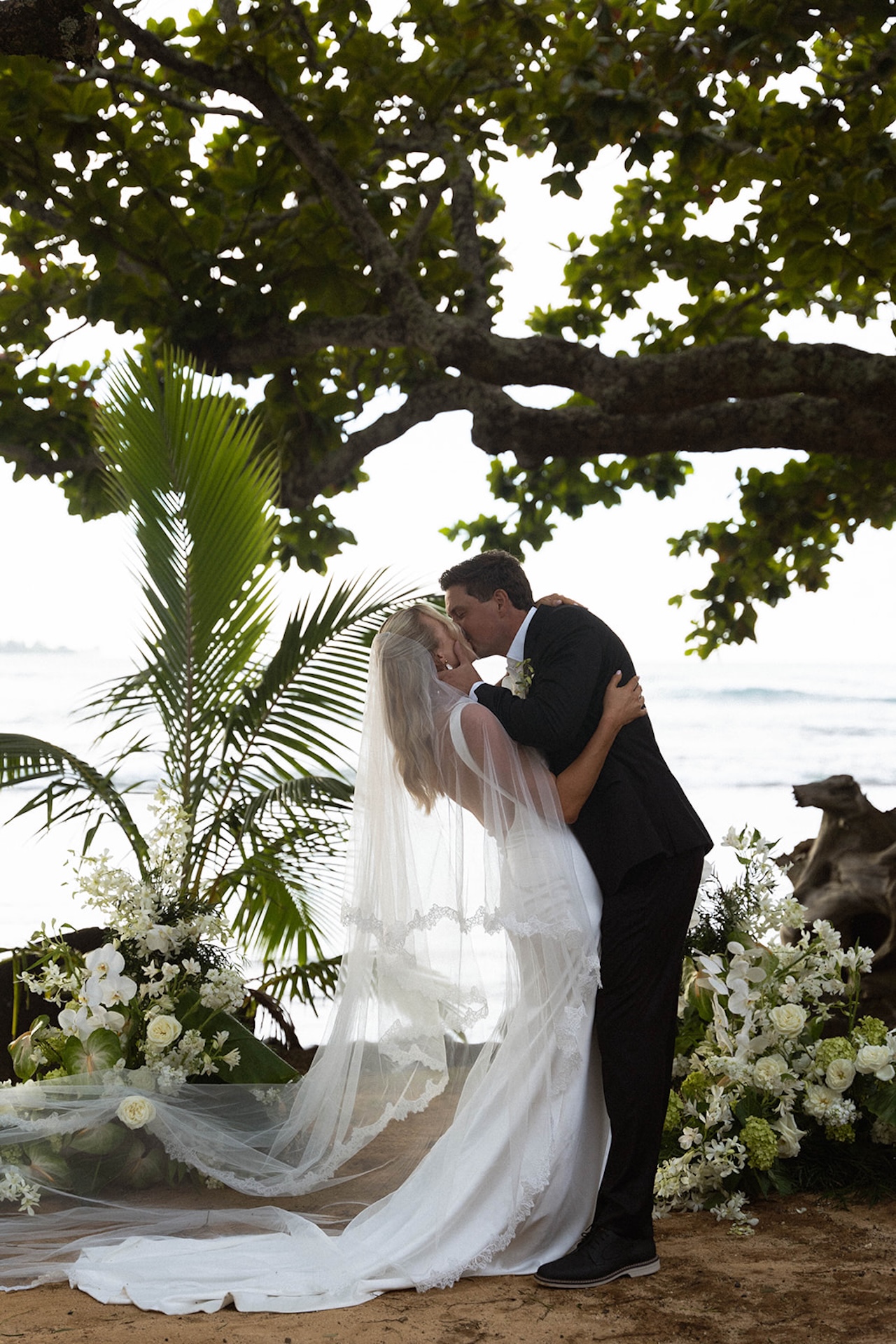 A bride and groom share a kiss beneath a large tropical tree overlooking the ocean during an intimate ceremony at one of the most scenic Kauai wedding venues.