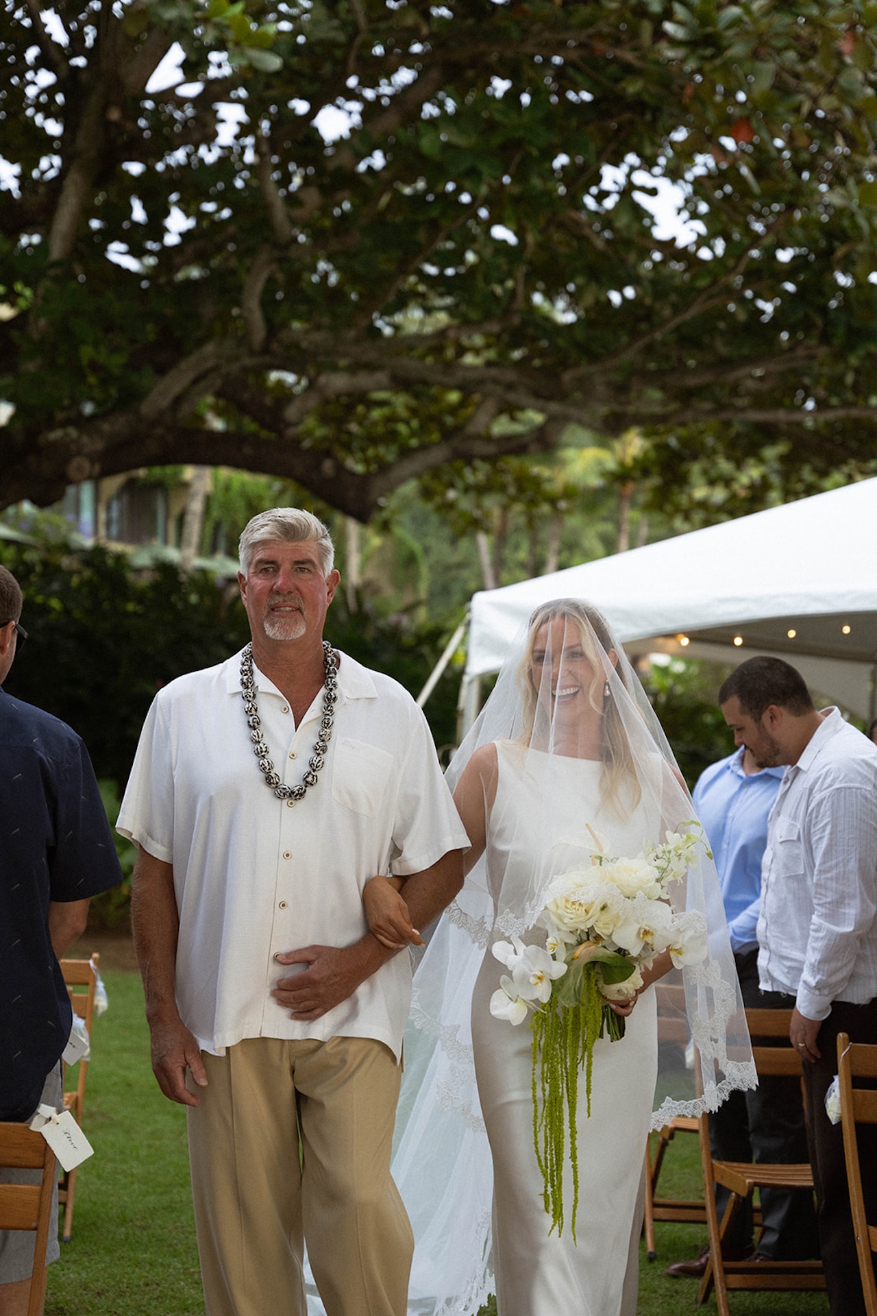 A bride walks down the aisle with her dad as guests look on, surrounded by tropical foliage and soft evening light.