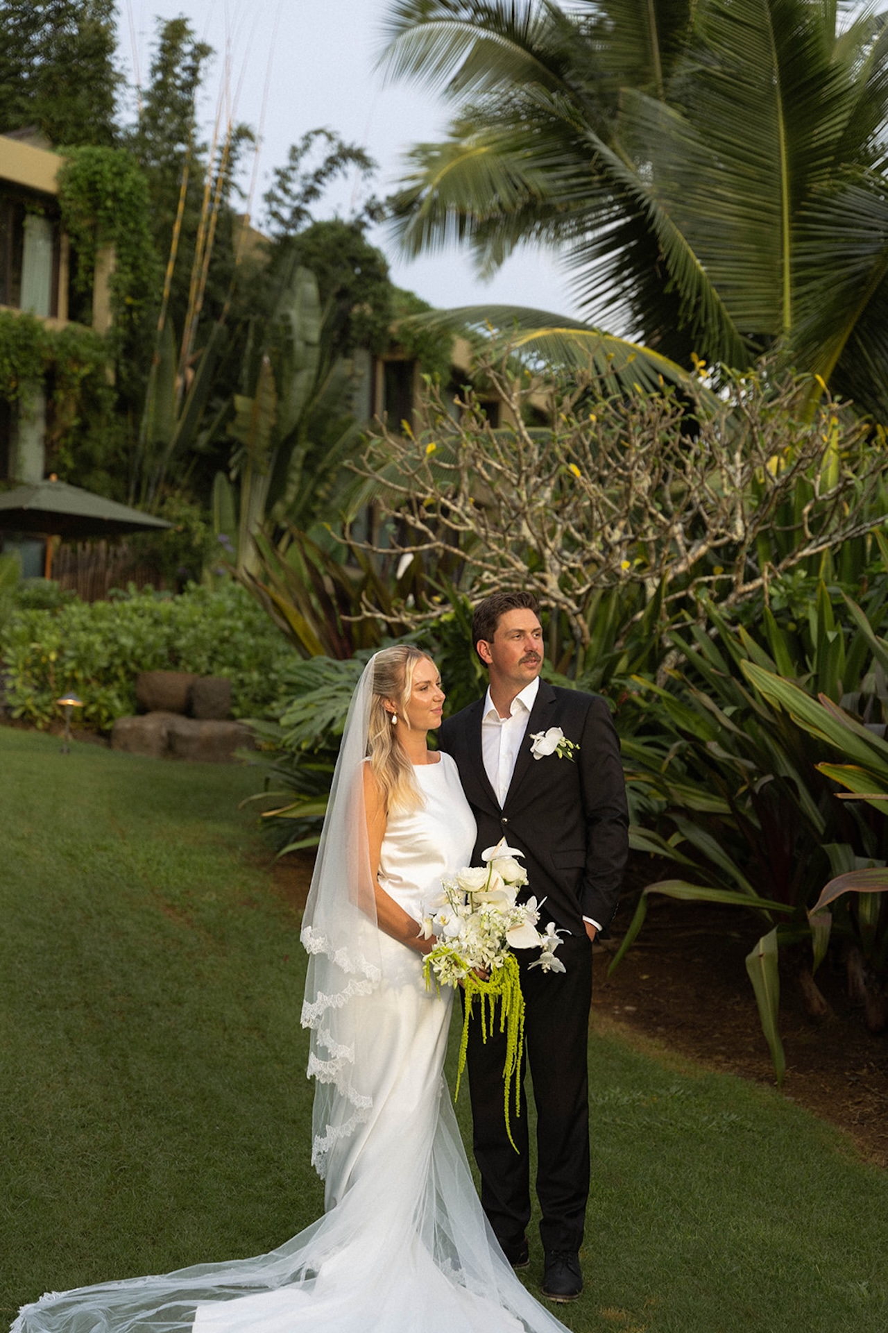 A bride and groom stand together on lush green grounds surrounded by tropical landscaping at one of the most picturesque Kauai wedding venues.