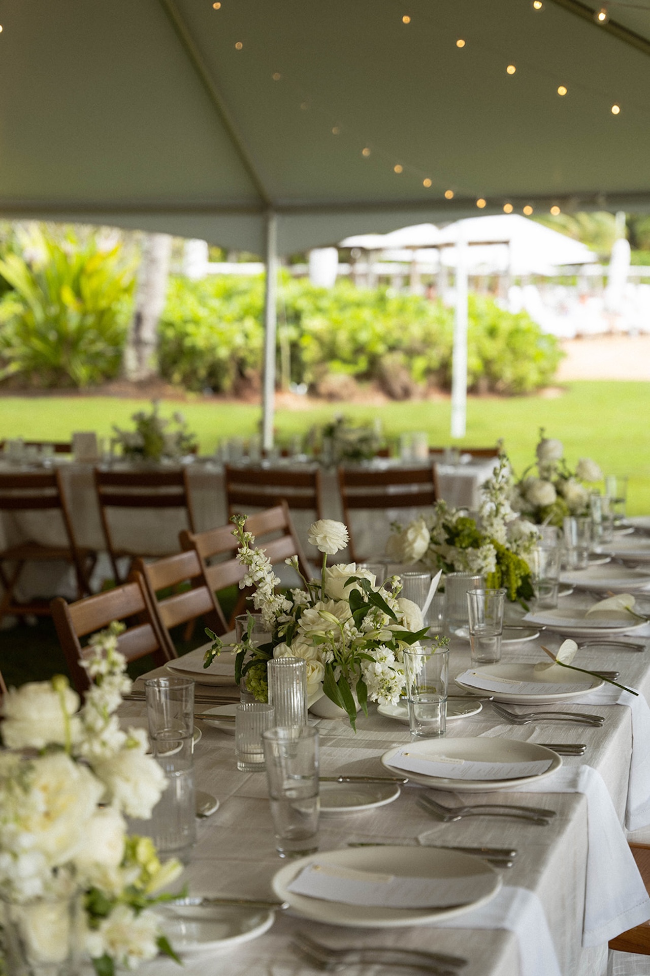 Long reception tables arranged beneath a sailcloth tent with wooden chairs, white florals, and string lights.