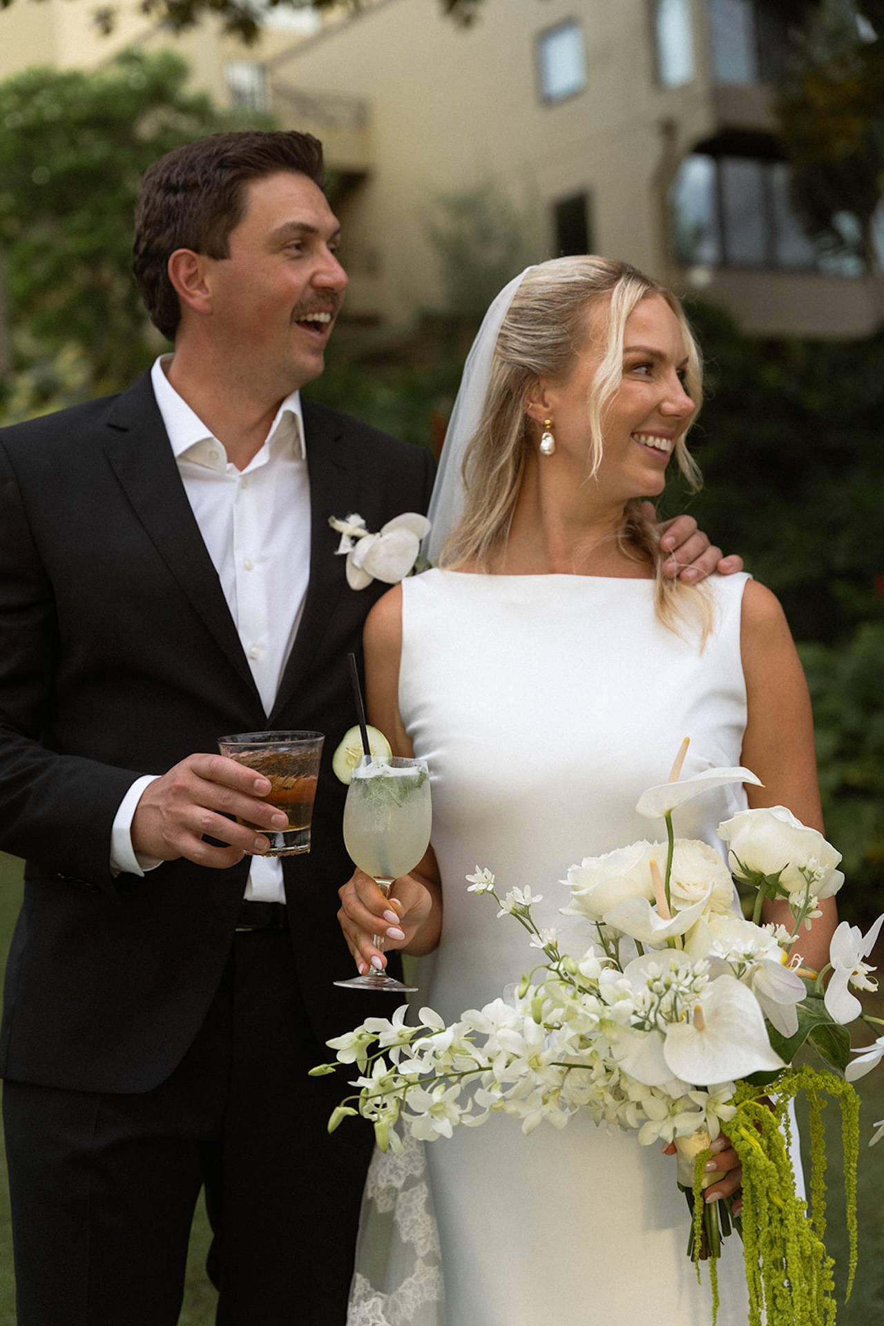 The bride and groom share drinks and laughter during cocktail hour, surrounded by greenery and white floral arrangements.