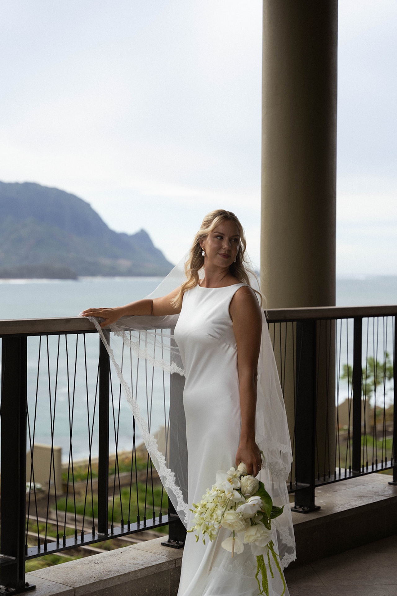 A bride stands on a balcony overlooking the ocean and mountains, holding a white floral bouquet at one of the most scenic Kauai wedding venues.