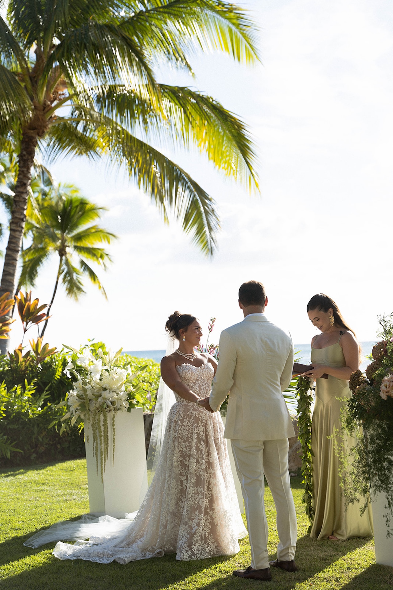 Bride and groom holding hands at their outdoor ceremony beneath palm trees.