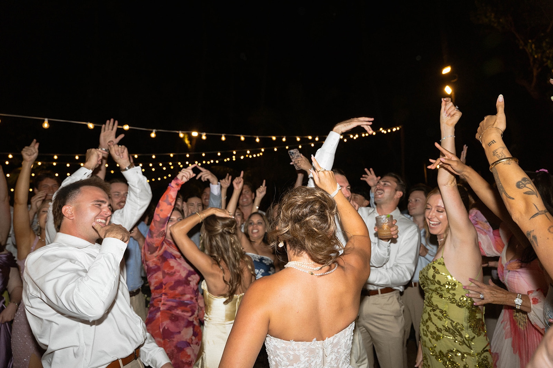 A large group of guests dances together beneath string lights at the Lanikuhonua wedding.