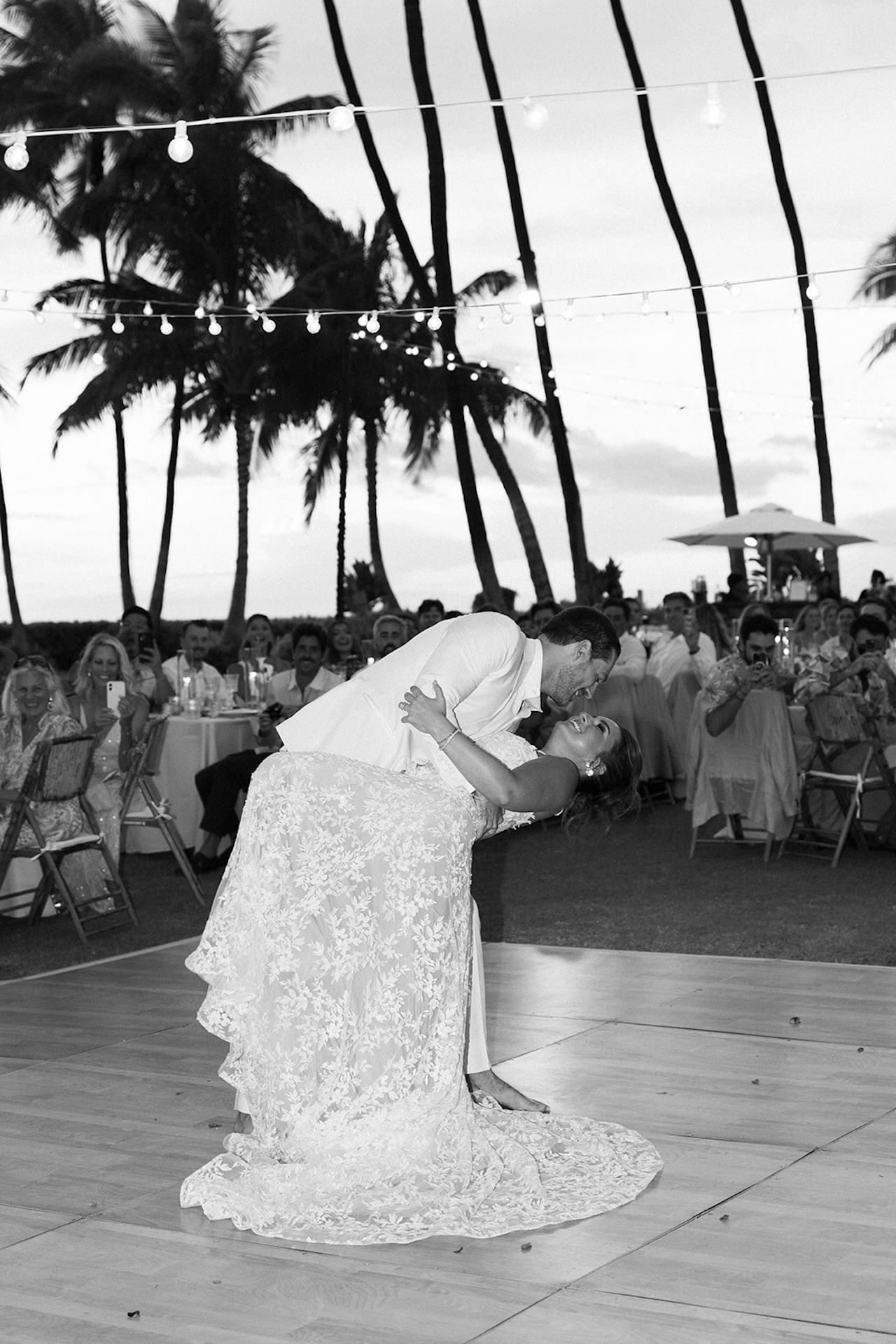 The groom dips the bride during their first dance beneath string lights at sunset.