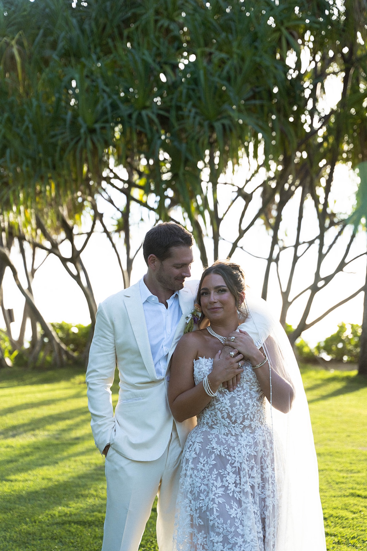 The groom wraps his arm around the bride as they stand together under the trees.
