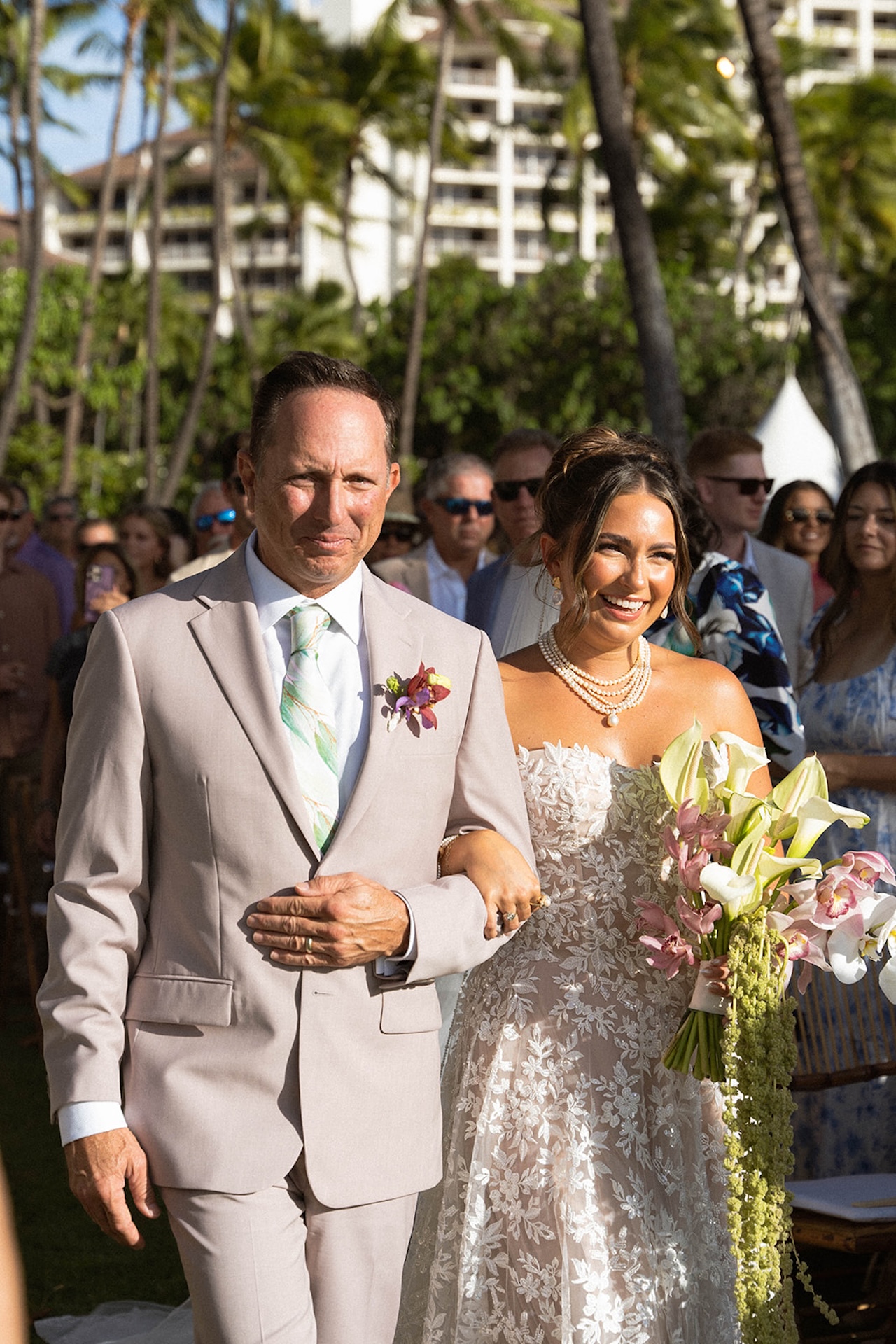 The bride walks down the aisle smiling beside her father in the afternoon sun.