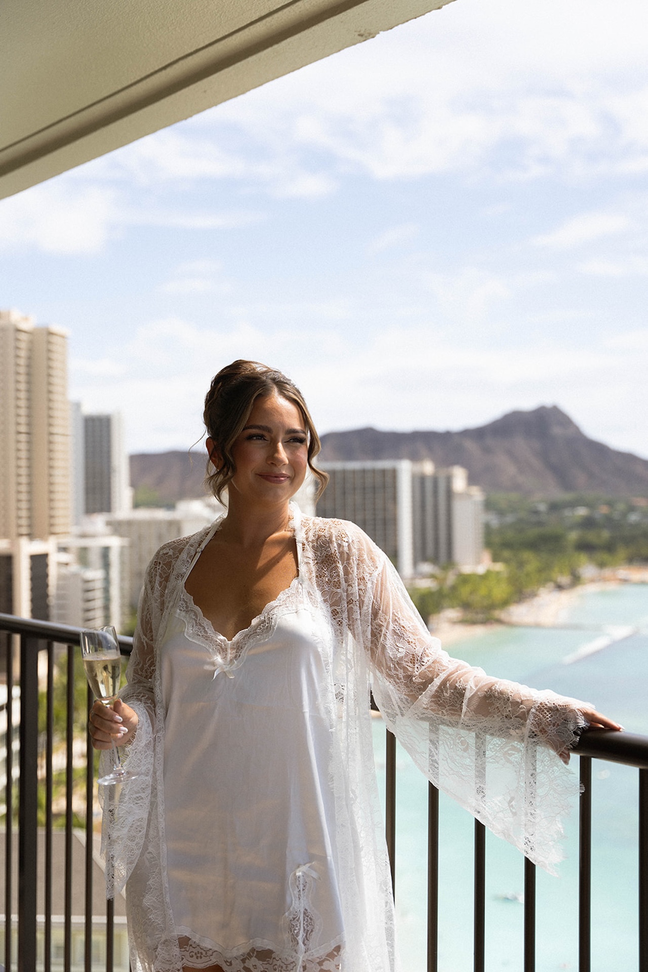 Bride in a lace robe stands on a balcony overlooking Waikiki before her Lanikuhonua wedding day.