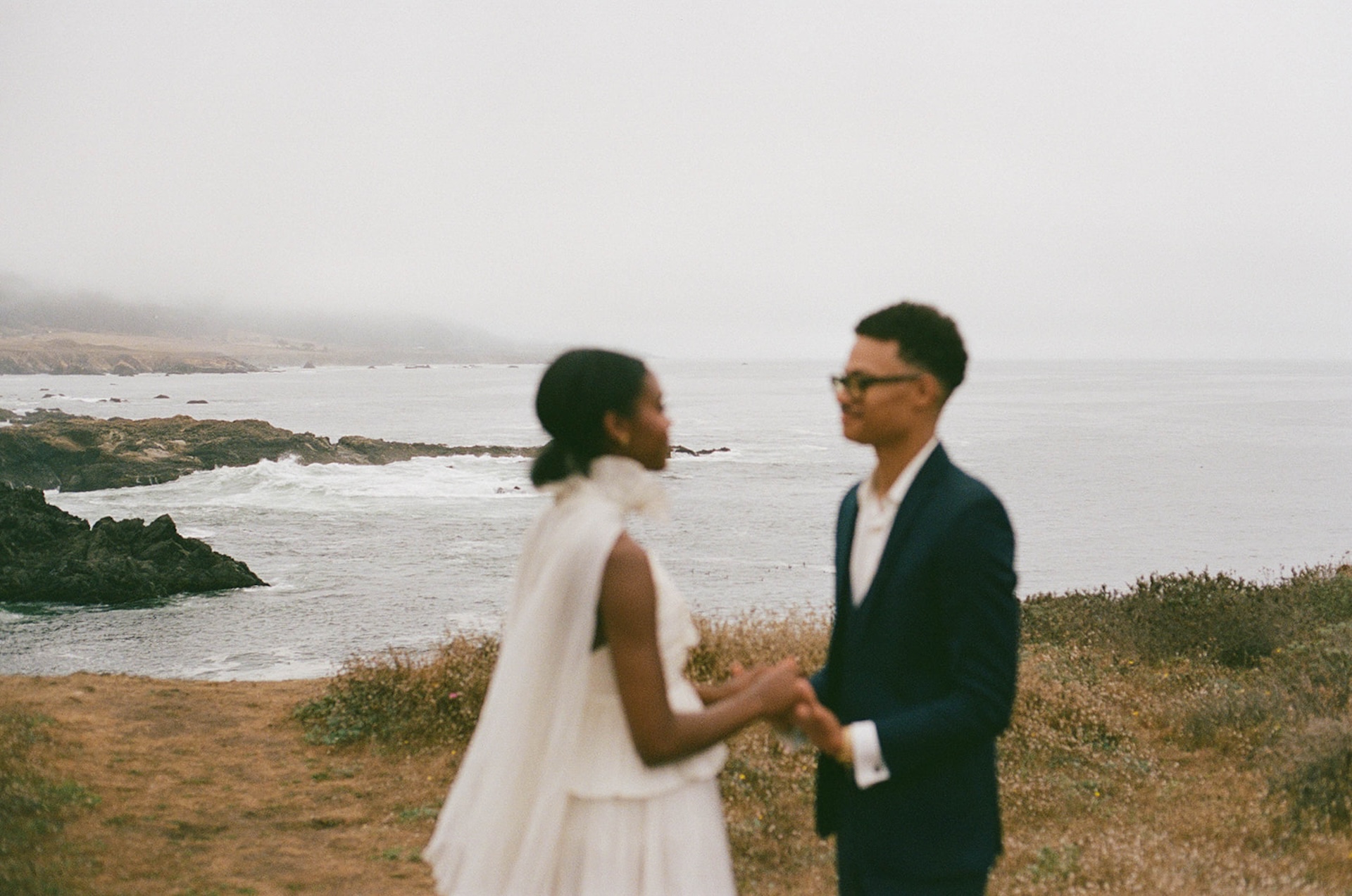 A stunning horizontal image of an out of focus bride and groom holding hands with the coastal cliffs in focus behind them during their Sea Ranch Lodge Wedding
