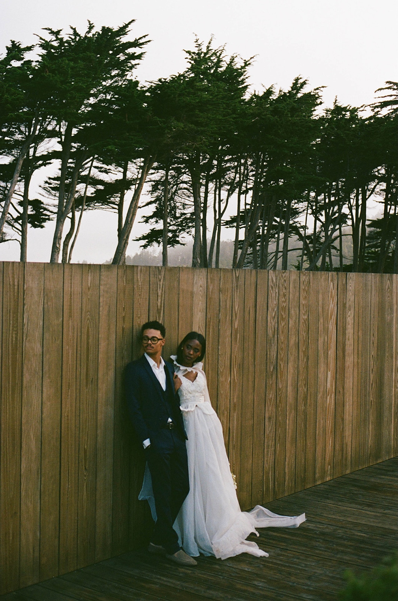 The bride and groom leaning against a wooden fence with trees and fog behind it during their bride and groom portraits.