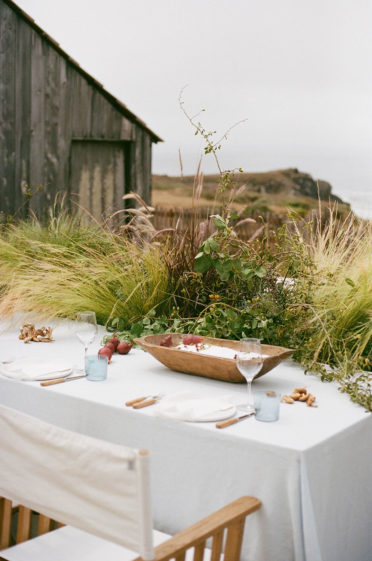 An elegant wedding reception table setup with grass and a barn in the background overlooking the California coast. The table has glass plates, blue cups, and a huge candle with florals around it.