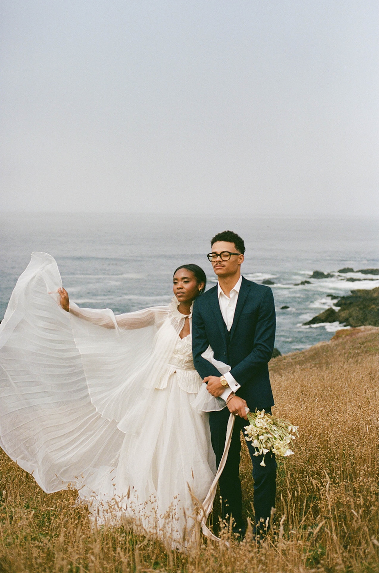 The bride and groom looking off into the distance and hugging onto each other with a stunning view of the California coastal cliffs behind them during their Sea Ranch Lodge Wedding