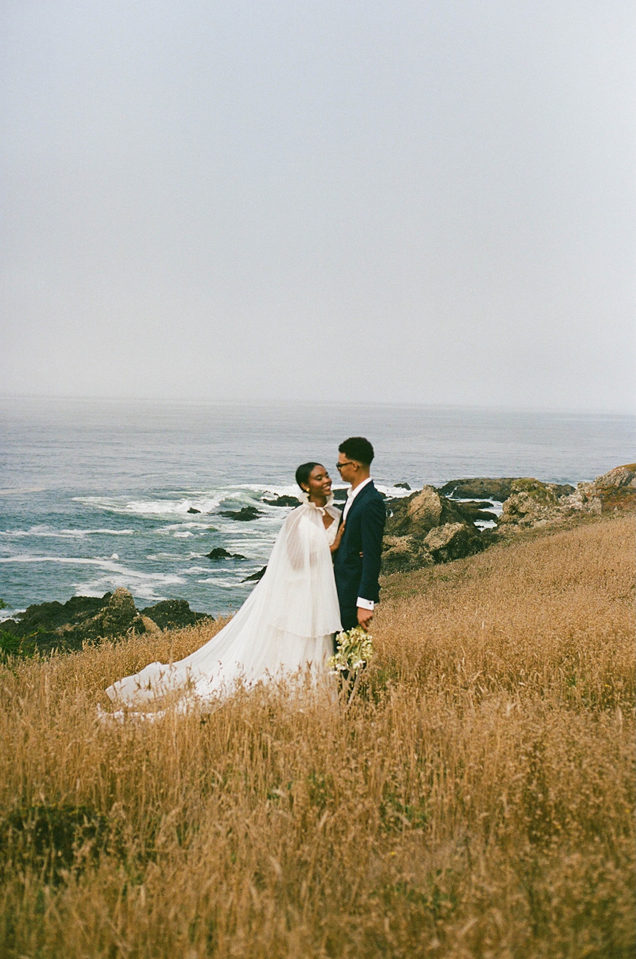 The bride and groom hugging and laughing at each other during their bride and groom portraits while standing in a grassy field as the waves crash against the cliffs on the California coast during their Sea Ranch Lodge Wedding