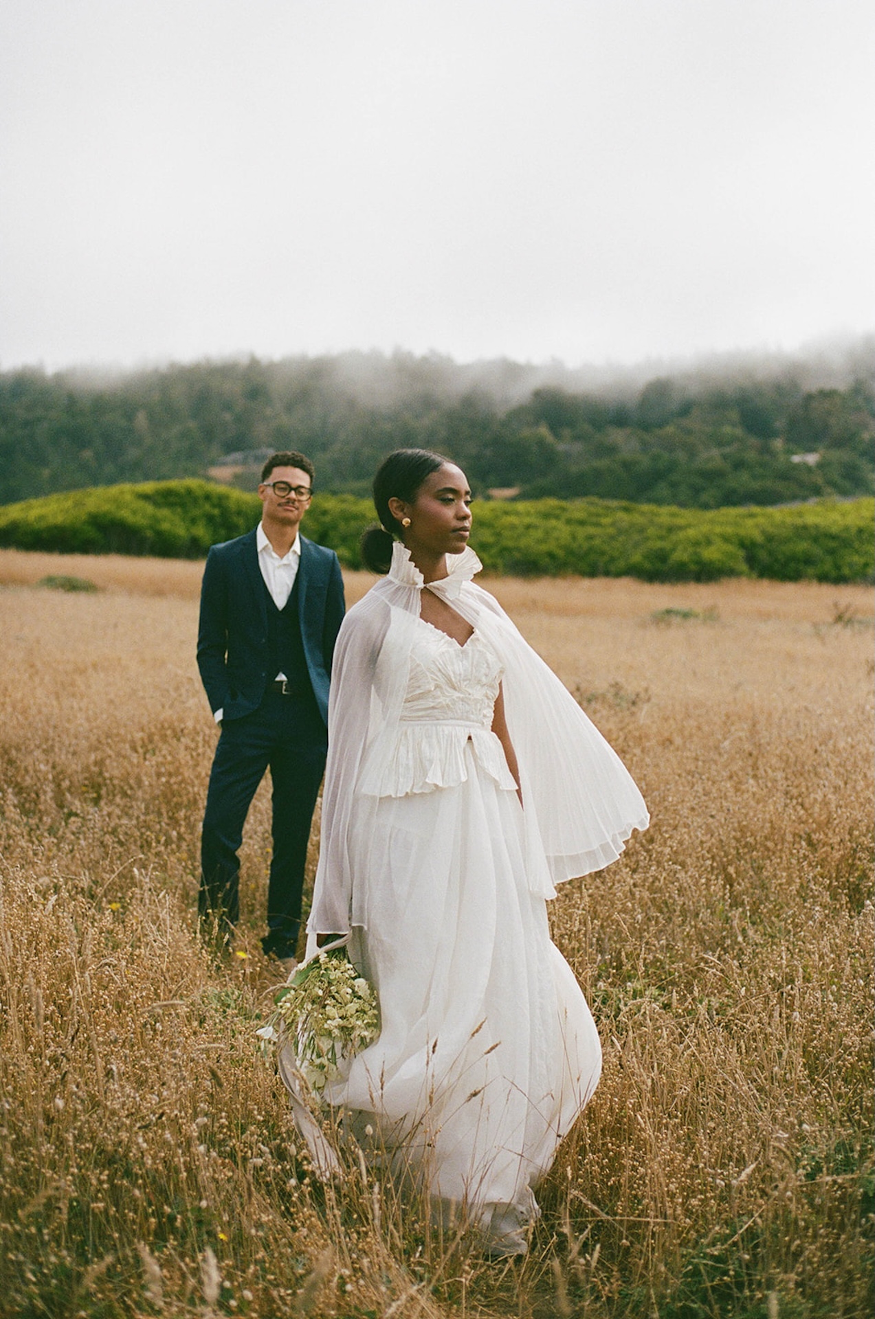 The bride standing in the foreground looking out at the water with the groom standing a couple steps back but blurred out as he looks at his bride. Foggy mountains and trees in the background.