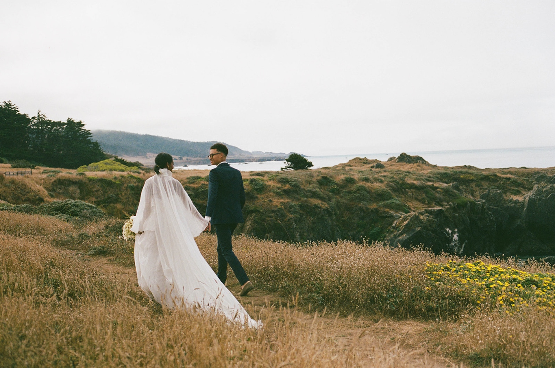 The bride and groom holding hands and walking on a dirt parth back to their Sea Ranch Lodge Wedding with the California cliffs in the background.