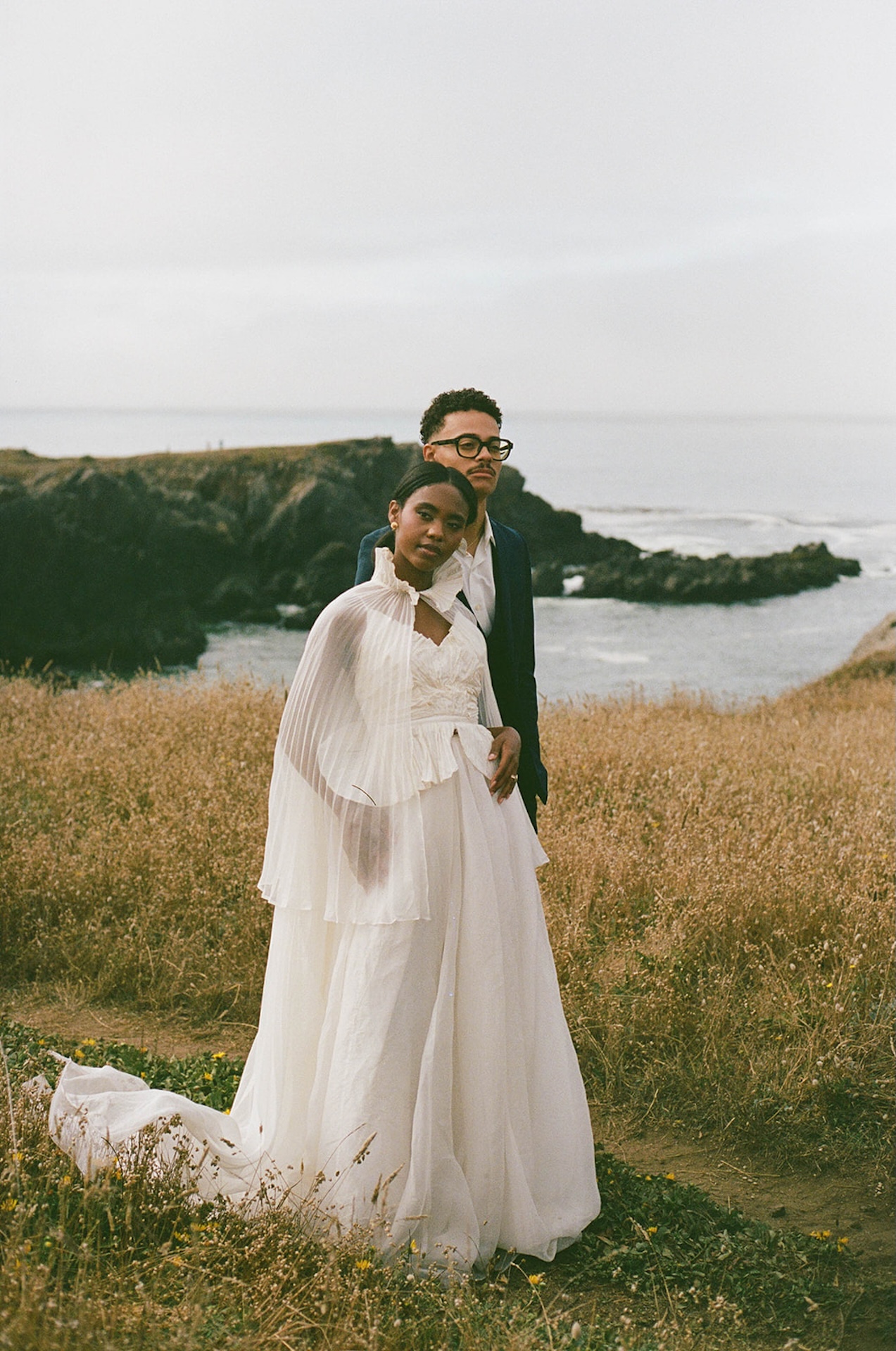 The bride and groom posing for an editorial photo on a dirt path. The groom is standing and looking out into the distance as the bride leans against him and looks towards the camera.
