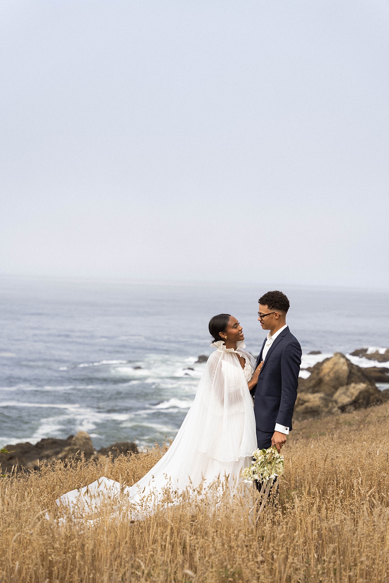 The bride and groom standing together in a grassy field overlooking the California coast during their couple portraits at their Sea Ranch Lodge Wedding
