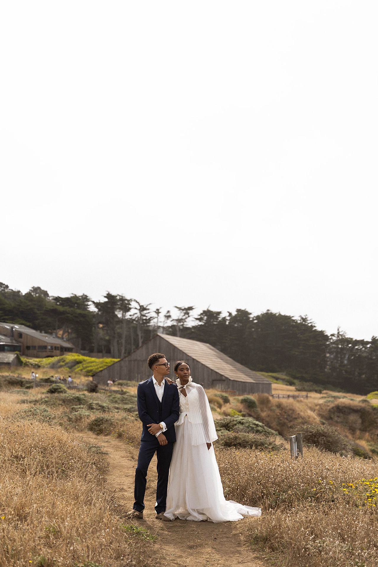 The bride and groom walking on a dirt path with the barn at Sea Ranch Lodge behind them as they look out at the water.