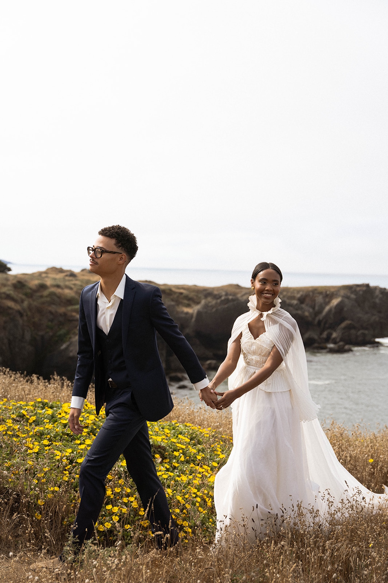 The bride and groom walking hand in hand in a field of yellow flowers and grass with coastal cliffs in the background during their Sea Ranch Lodge Wedding
