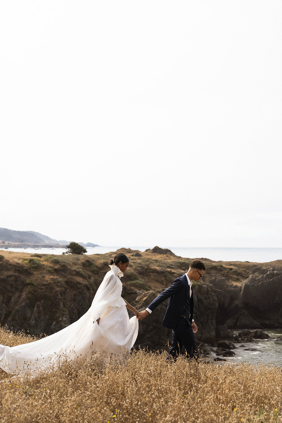 The bride and groom walking along the grassy cliffs during their bride and groom portraits for their Sea Ranch Lodge Wedding
