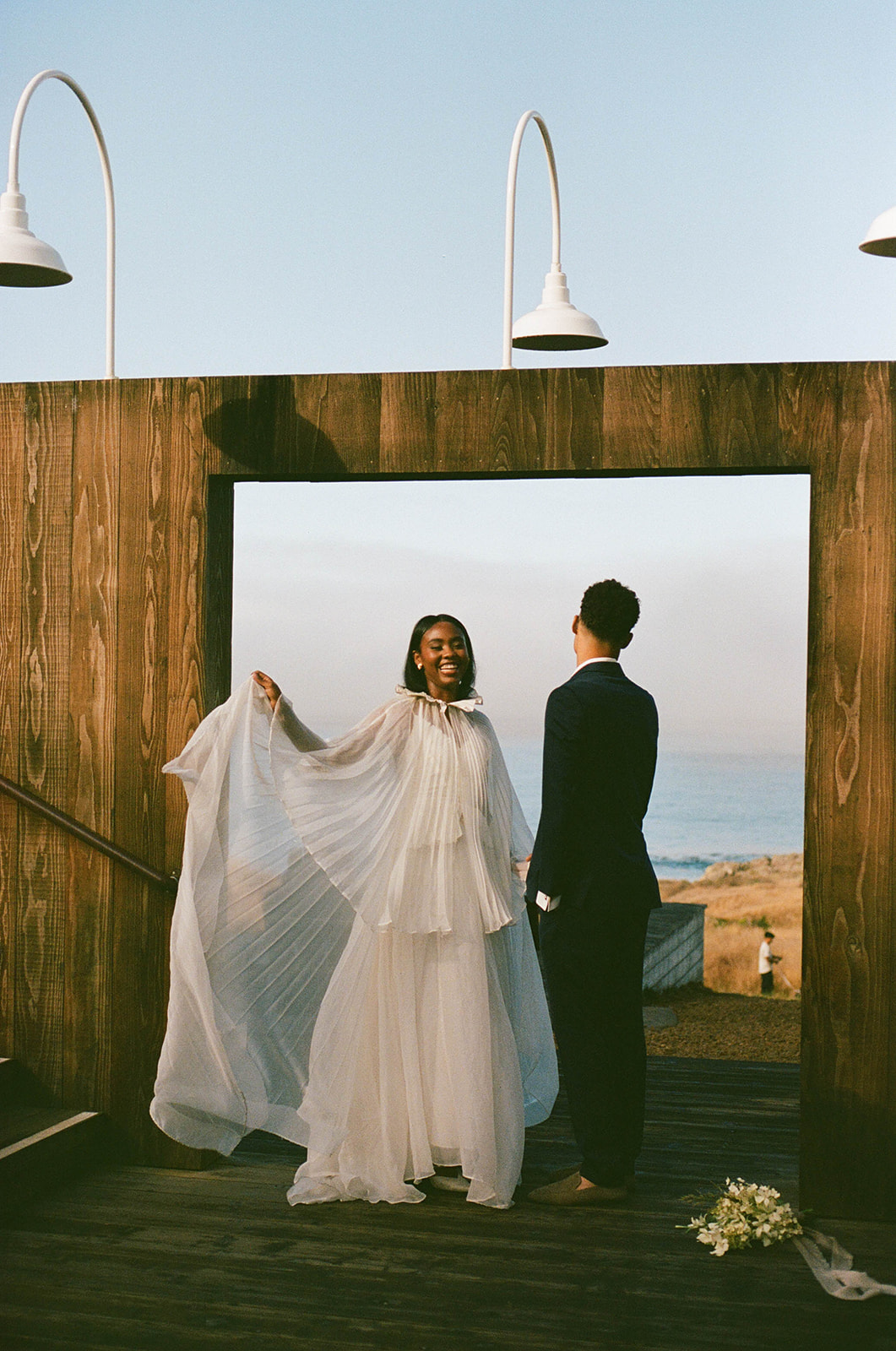 A bride playing with her dress in a wood doorway with the California coast in the background while the groom holds her hand and stands next to her during their Sea Ranch Lodge Wedding
