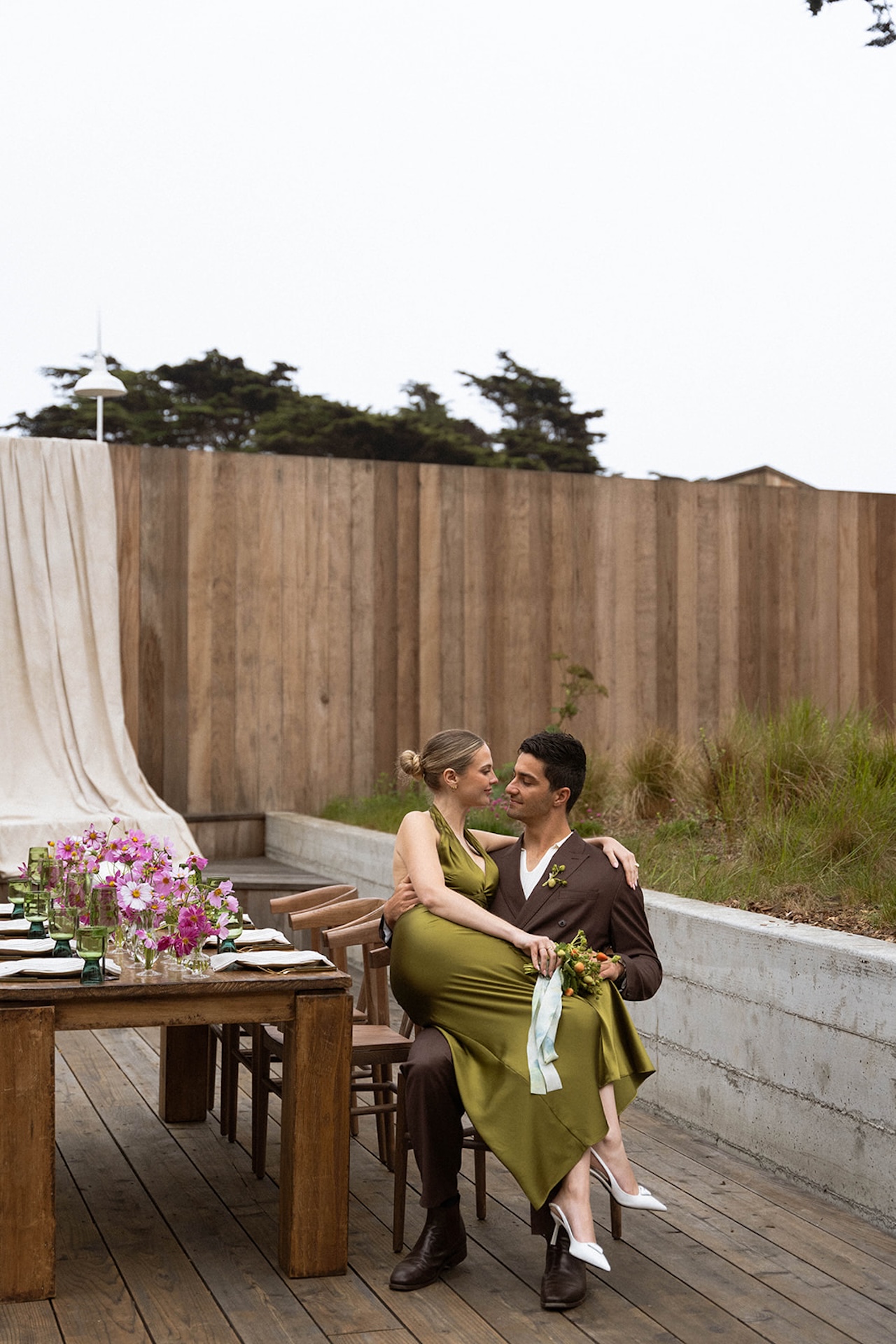 Bride sitting on the groom’s lap at an outdoor wedding table, both smiling and wearing modern olive and brown wedding outfits.
