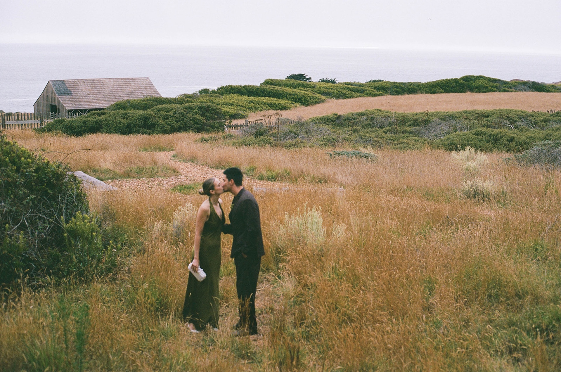 Wide editorial shot of bride and groom standing in an open coastal meadow with rolling hills and ocean views behind them.