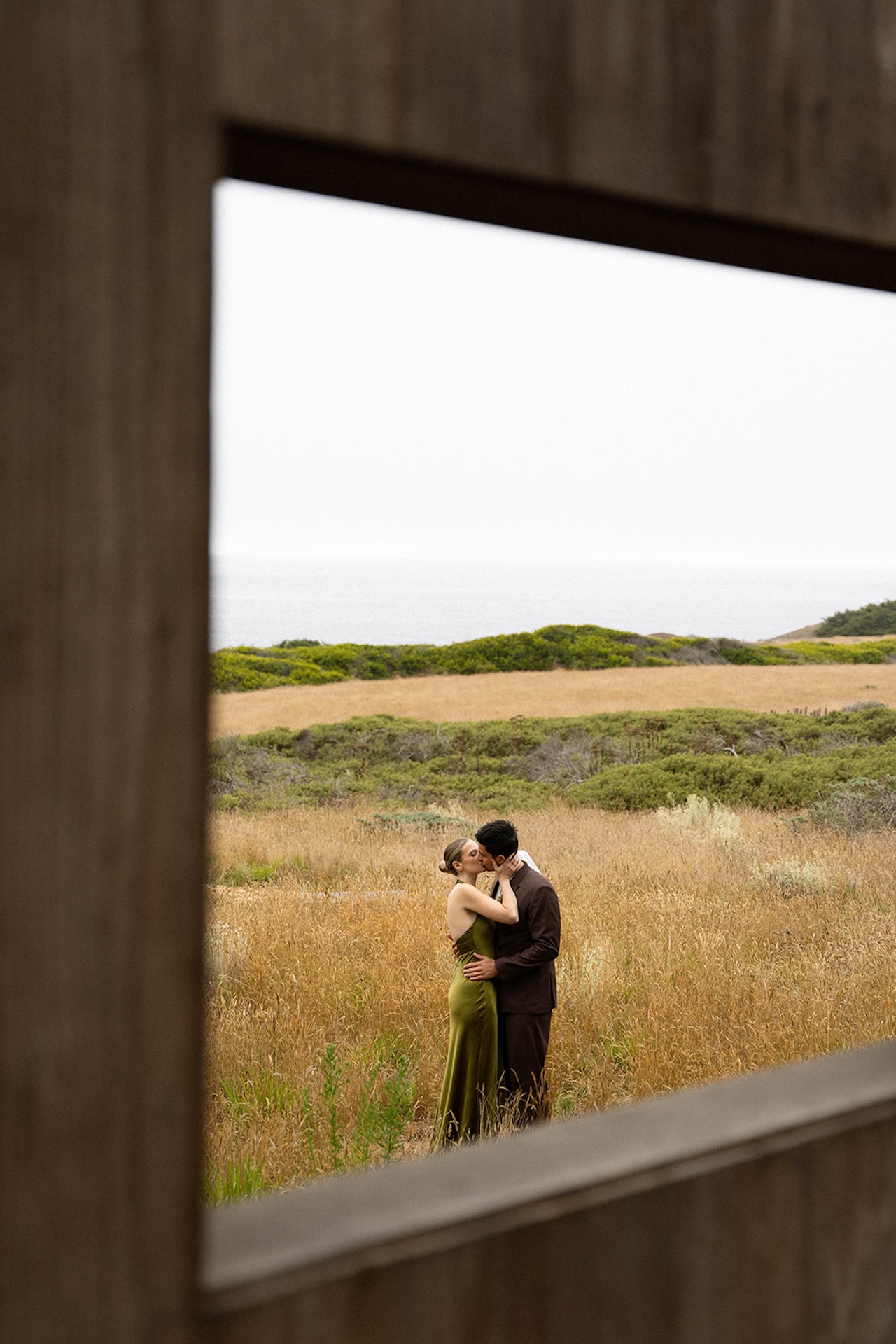 Bride and groom kissing in a coastal field with golden grass and soft ocean light in the background.