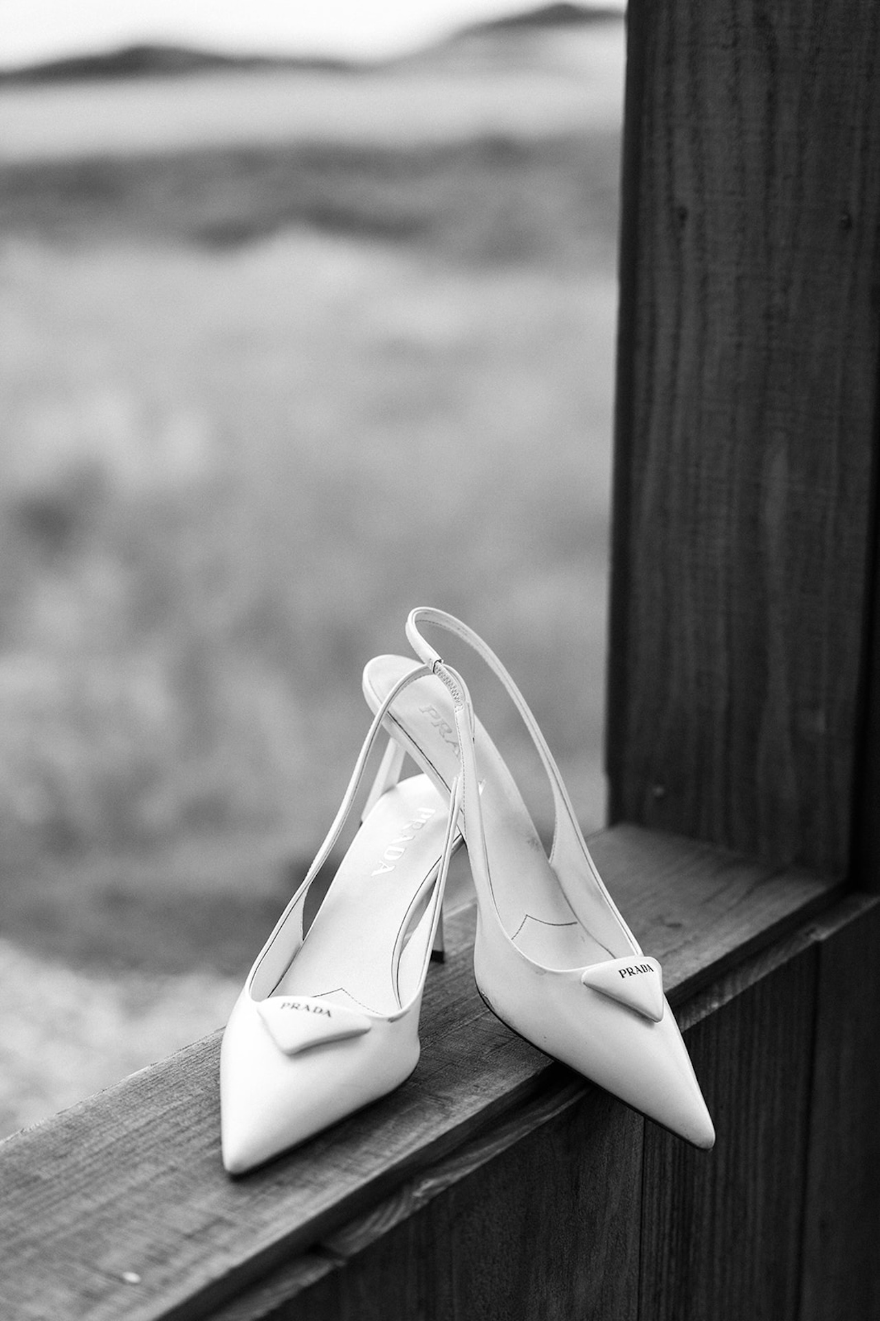Black and white photo of white Prada slingback heels resting on a wooden ledge overlooking a coastal landscape.