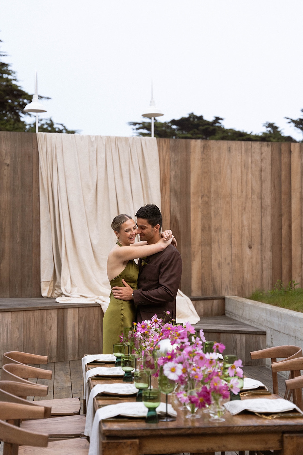 Bride and groom embracing beside a styled reception table with pink florals and green goblets at an outdoor coastal wedding.
