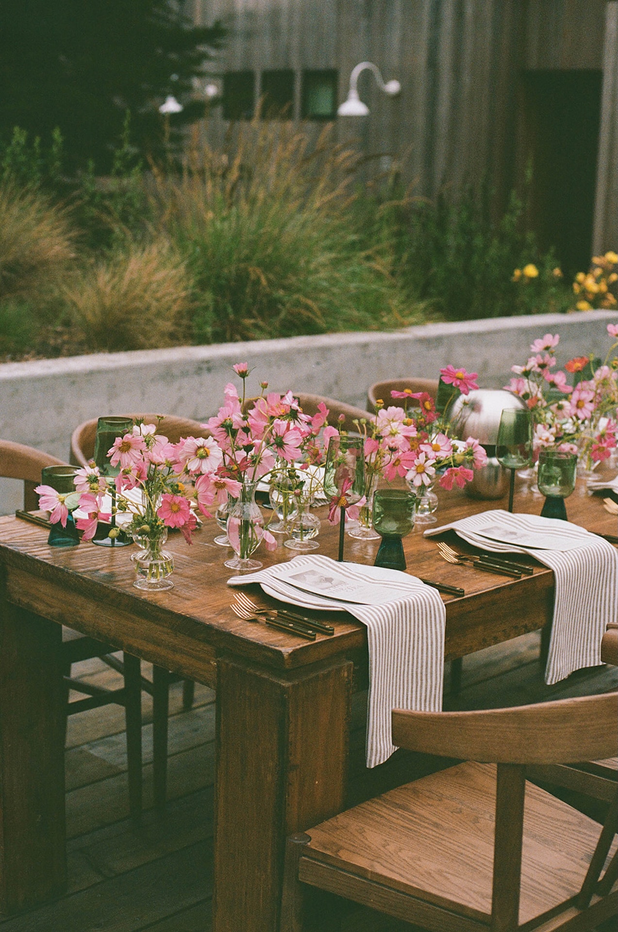 Outdoor wedding tablescape with wooden table, green glassware, striped linen runners, and pink floral arrangements in glass vases.