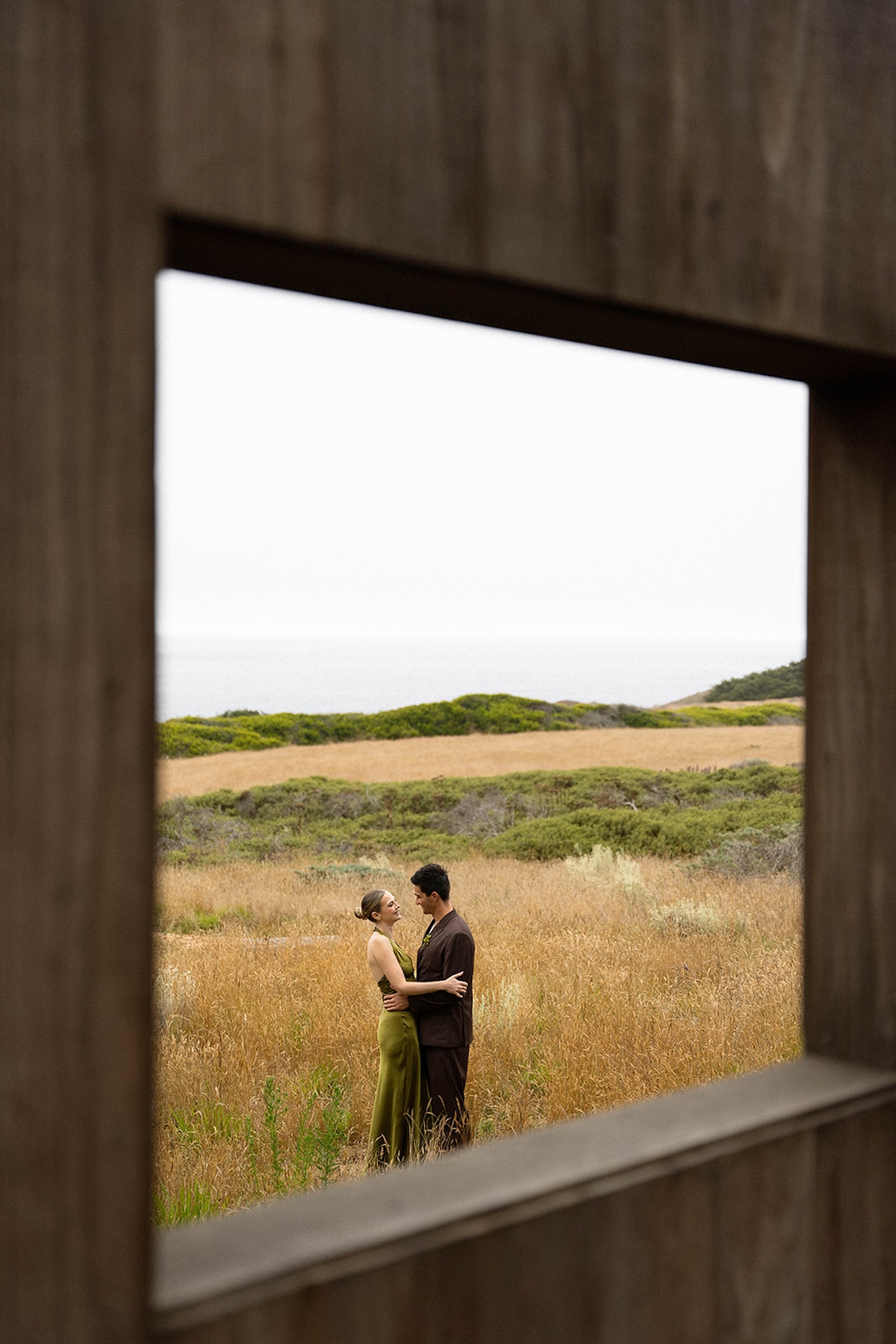 Bride and groom standing together in tall coastal grass, framed through a wooden window opening with the ocean in the distance.