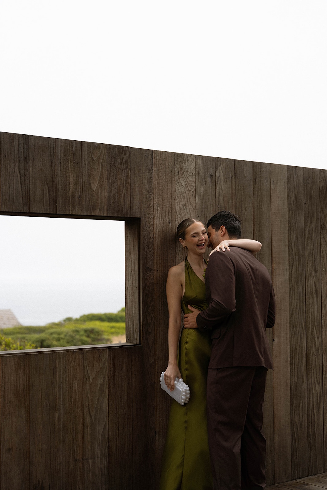 Bride laughing as the groom holds her close beside a wooden wall, capturing genuine connection while planning a destination wedding.