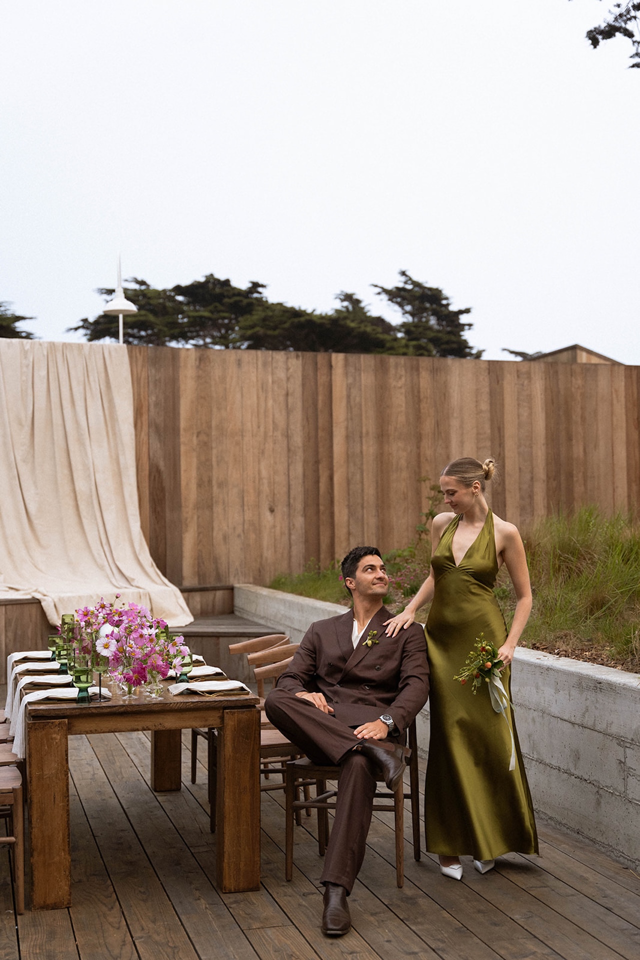 Bride standing beside the groom seated at a styled outdoor table, both exchanging a quiet look in a modern coastal setting.