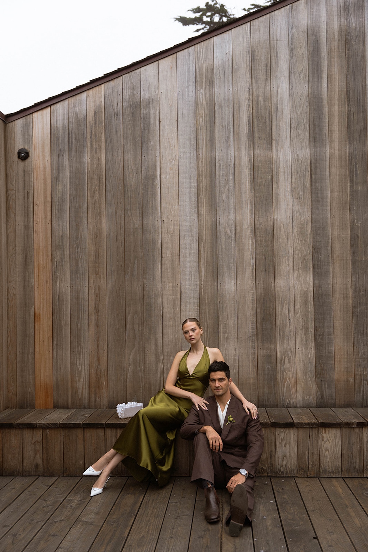 Editorial portrait of the couple seated against a wooden wall, blending relaxed posture with refined wedding styling.