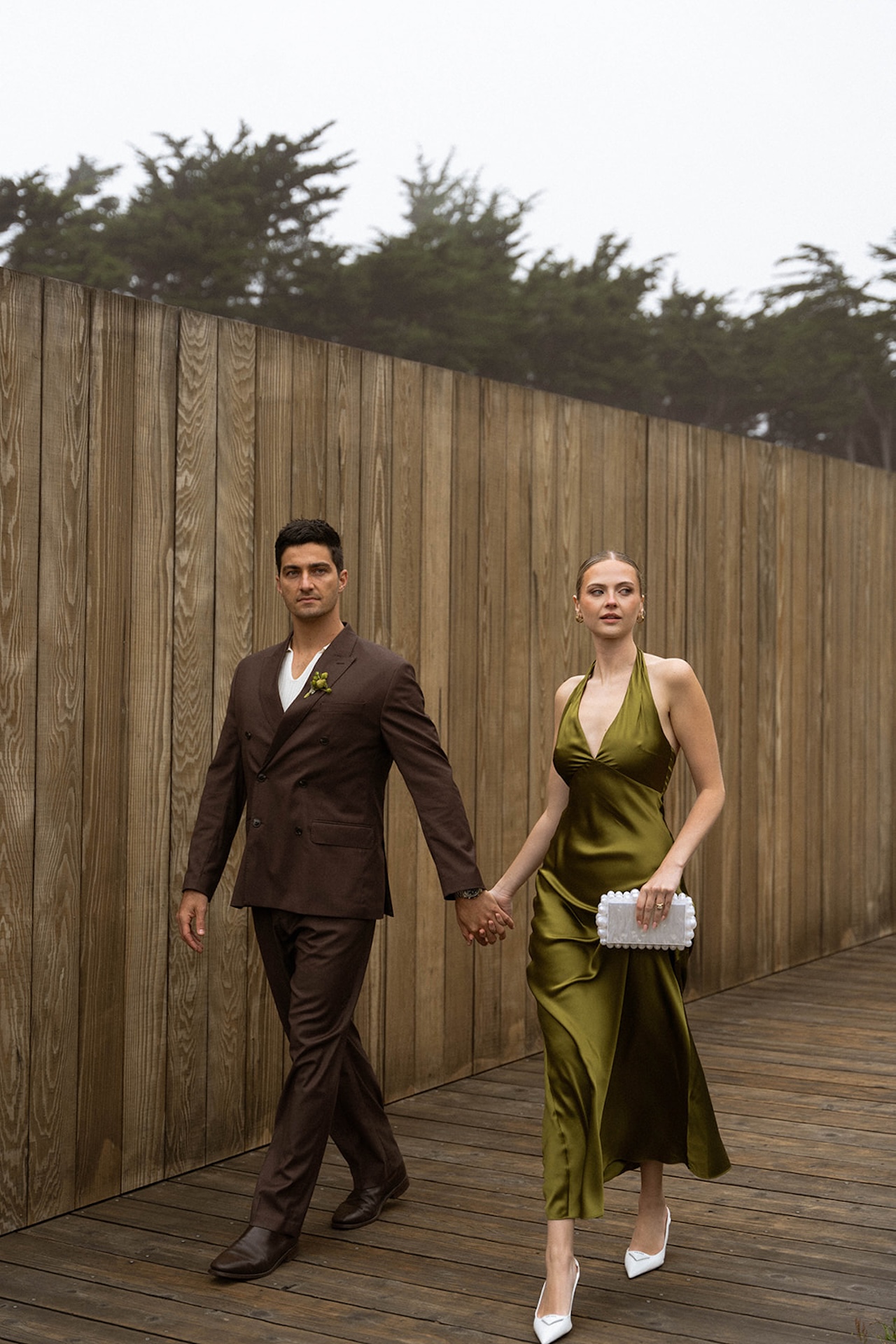 Couple walking on a wooden boardwalk as the groom lifts the bride, surrounded by coastal grasses and tall trees.