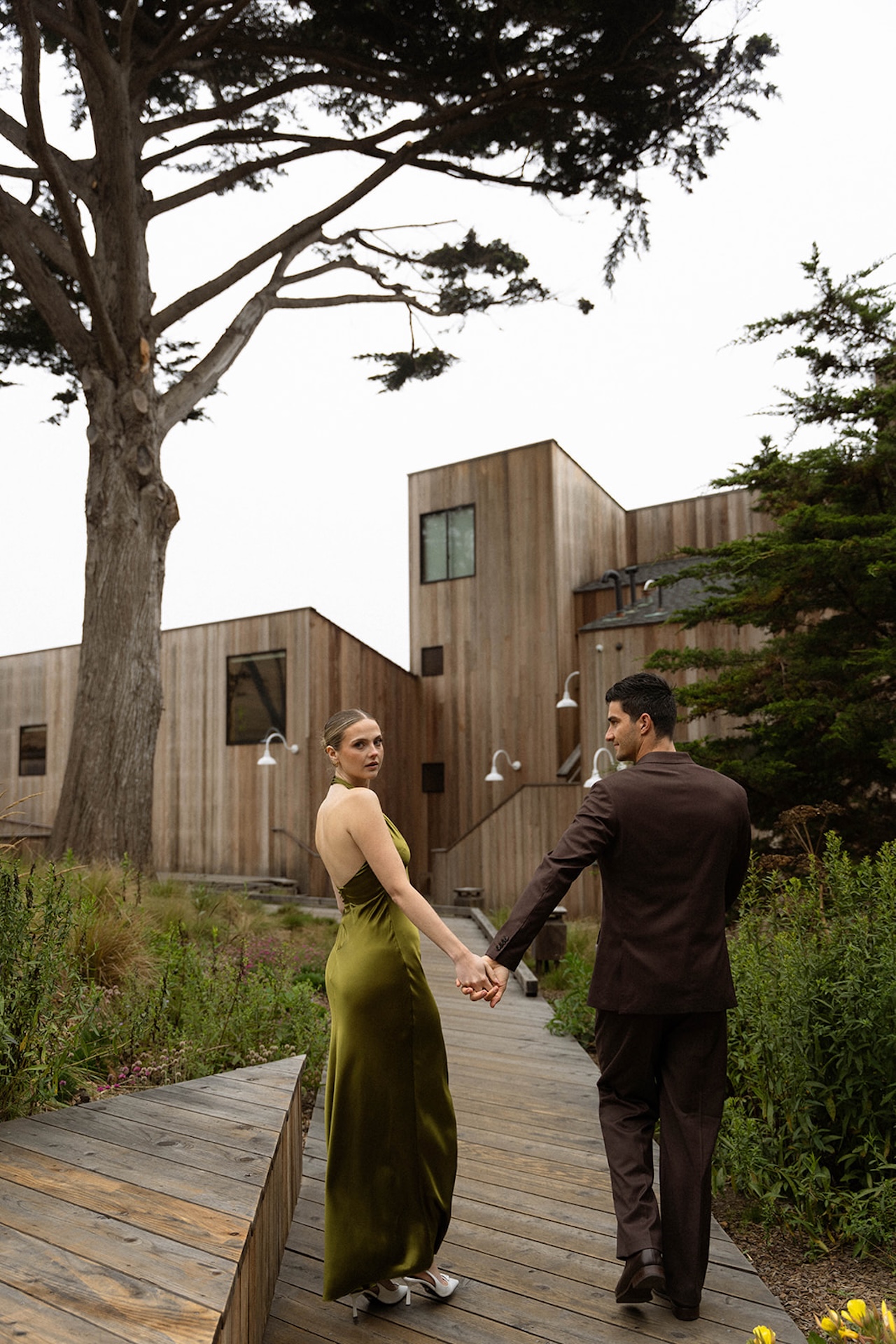Bride and groom walking away hand in hand through landscaped paths between wooden buildings, ending the day with quiet connection.