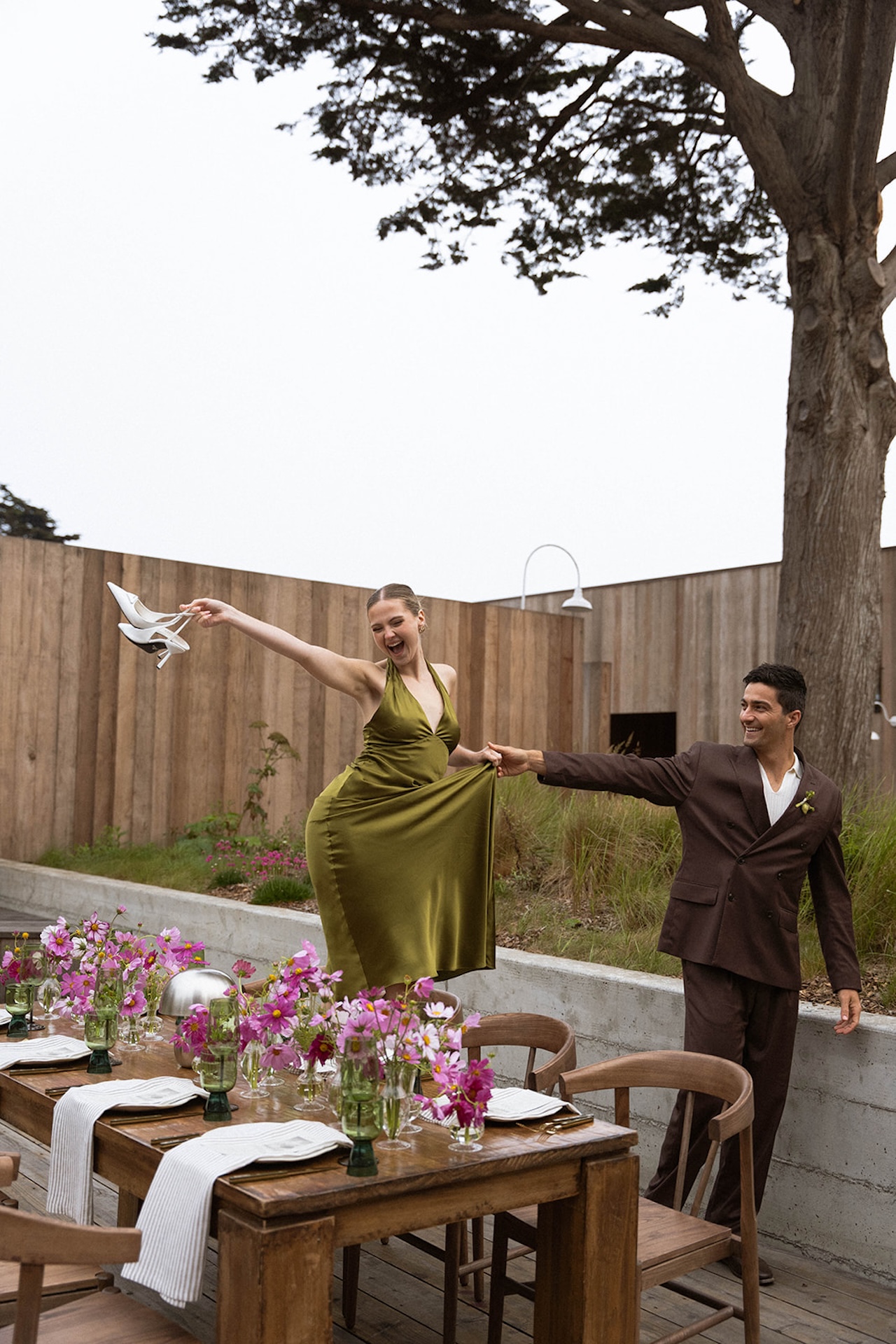 Bride playfully lifting her heels while holding the groom’s hand above a styled outdoor reception table.