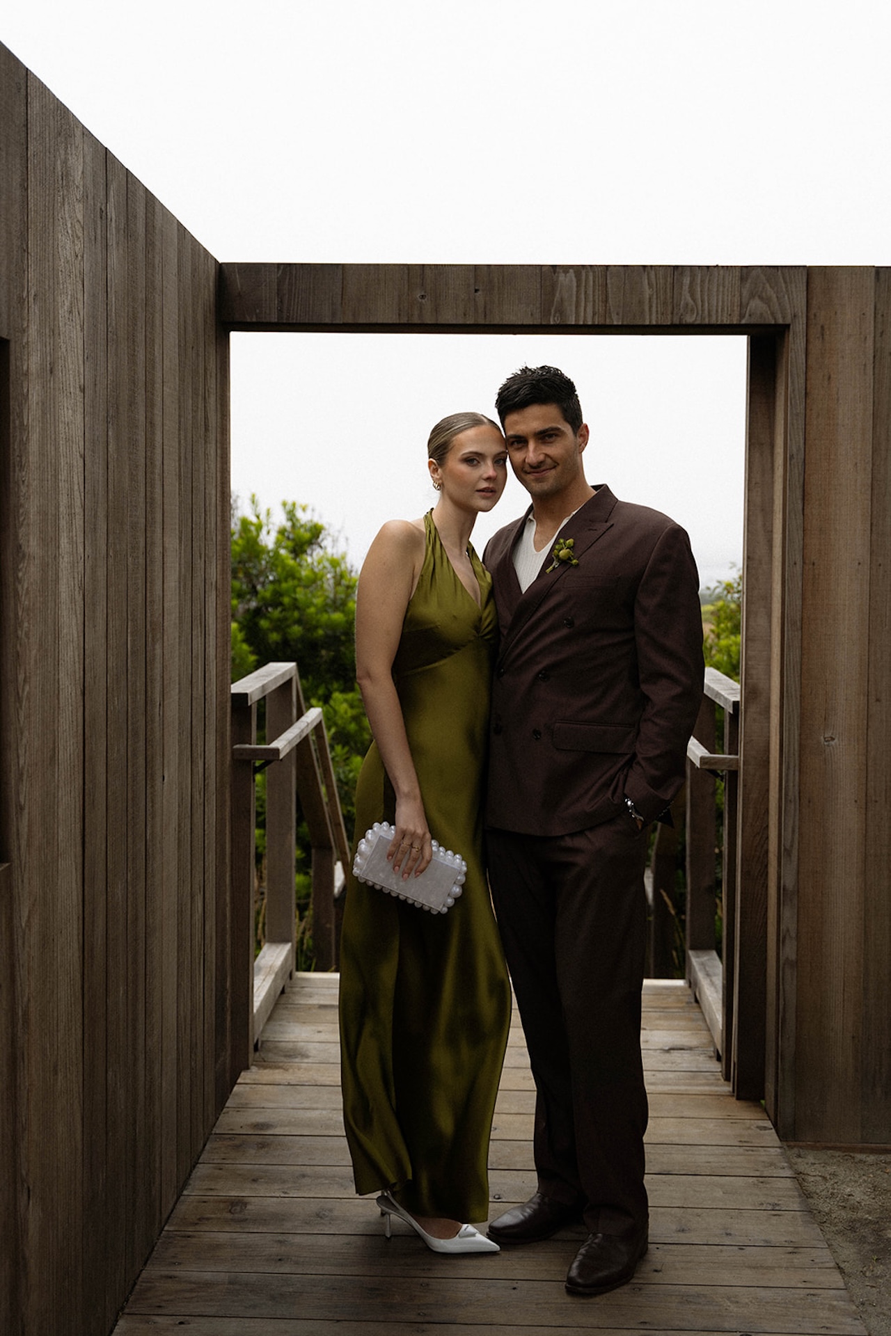Bride and groom standing together in a wooden doorway, styled in modern olive and brown attire while planning a destination wedding.