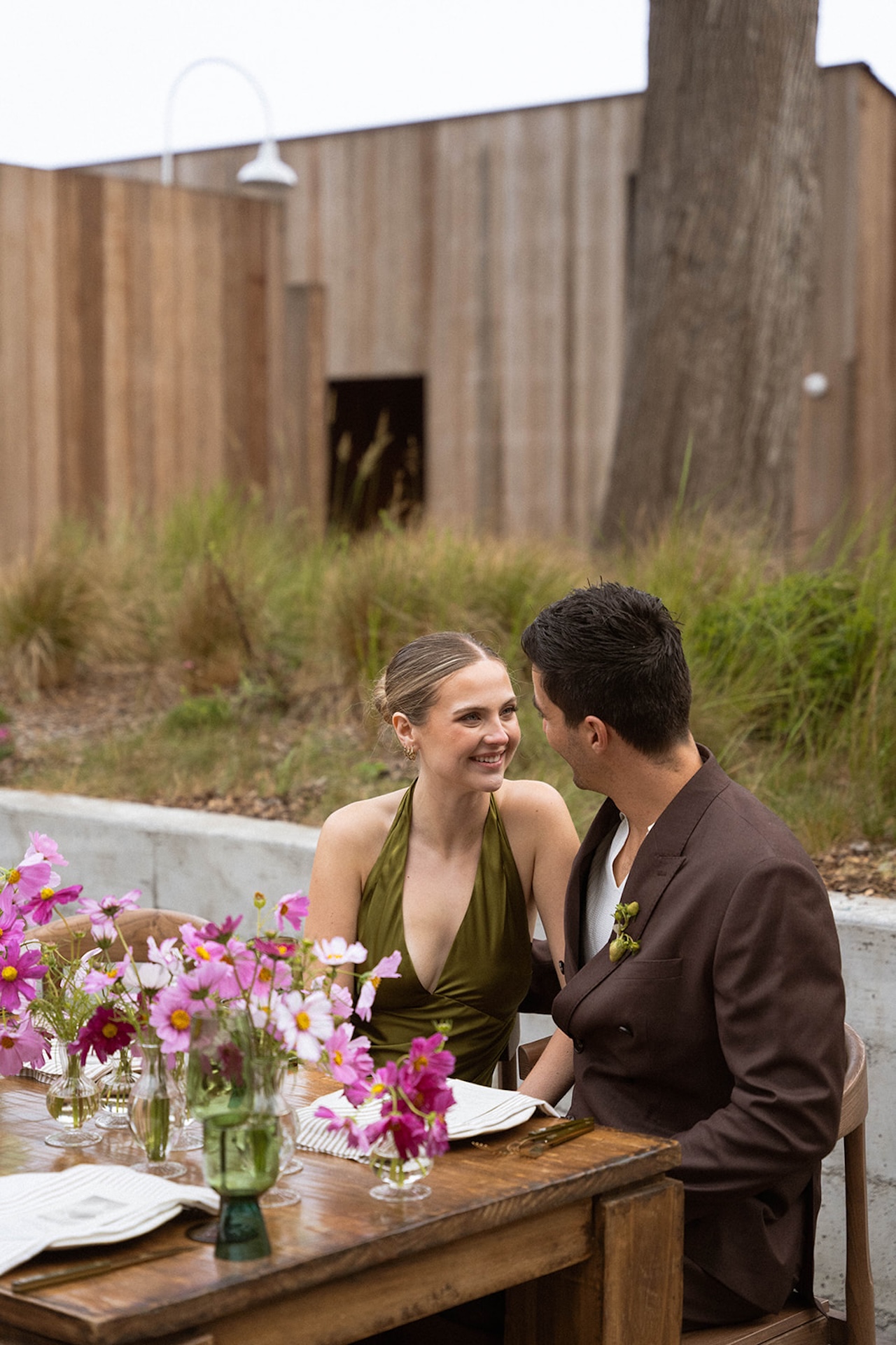 Couple sitting at an outdoor wedding tablescape with pink cosmos flowers, green glassware, striped linens, and wooden chairs styled for an intimate celebration while planning a destination wedding.
