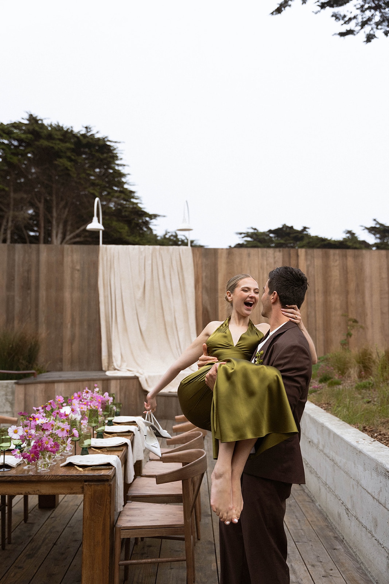 Bride and groom laughing as the groom carries the bride along a wooden path between modern cabins, ending the day with playful energy.
