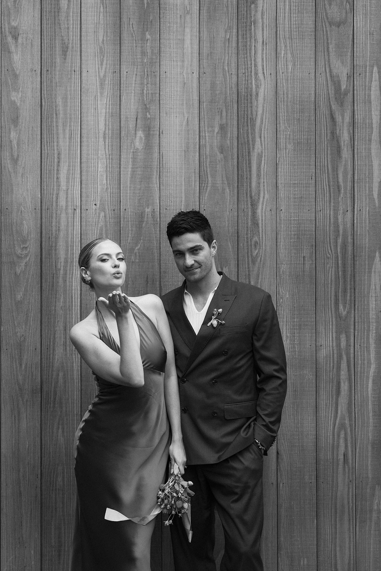 Black and white portrait of the couple standing together against a wooden backdrop, blending classic style with modern wedding fashion.