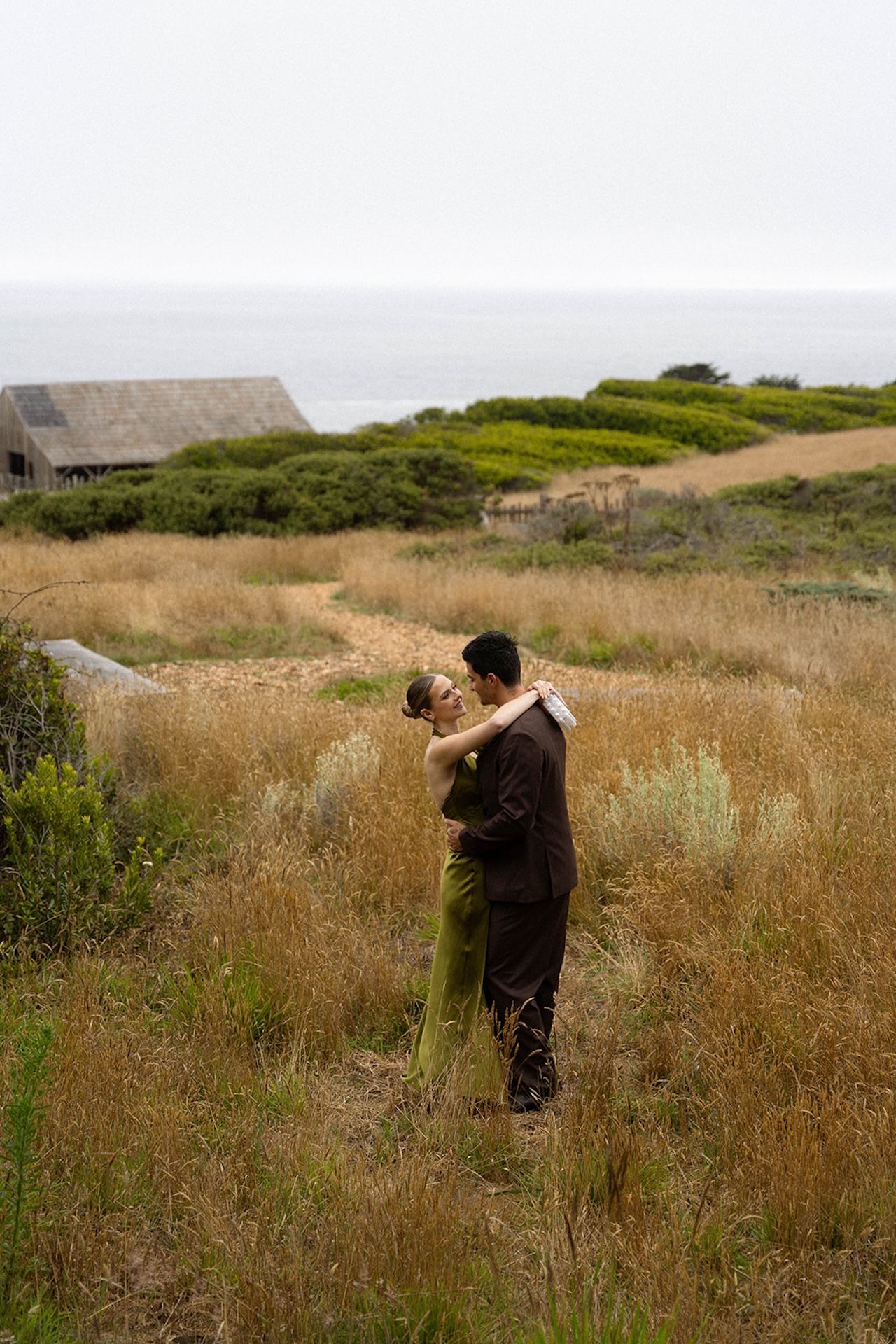 Couple embracing in tall coastal grass with the ocean in the distance, styled in rich tones that complement the natural landscape.